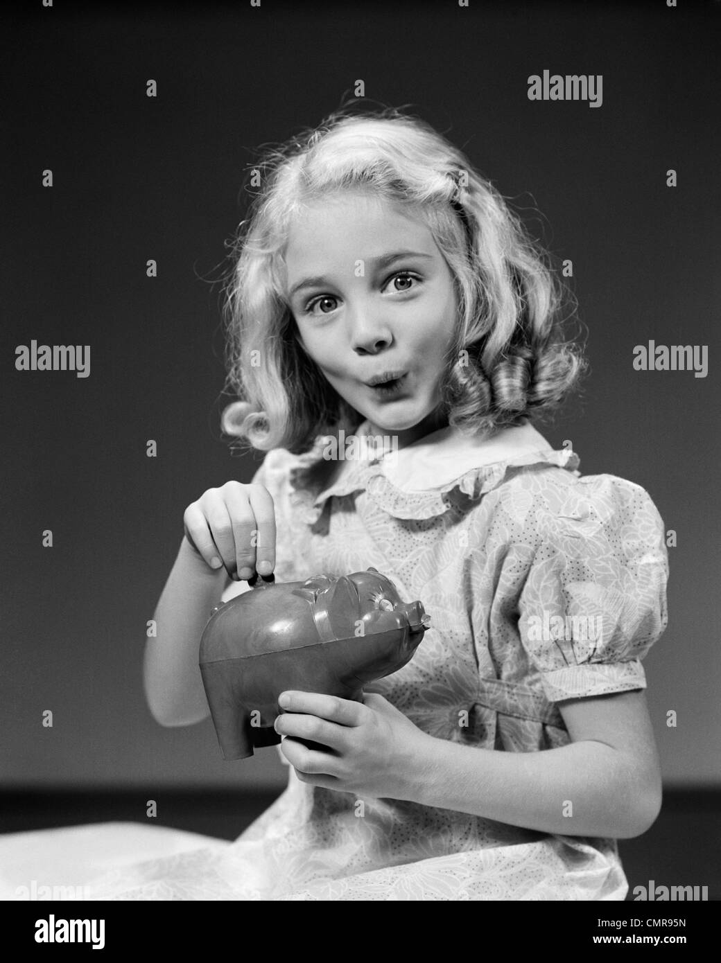 1940s CHILD BLOND GIRL PUTTING MONEY COIN INTO PIGGY BANK LOOKING AT CAMERA Stock Photo