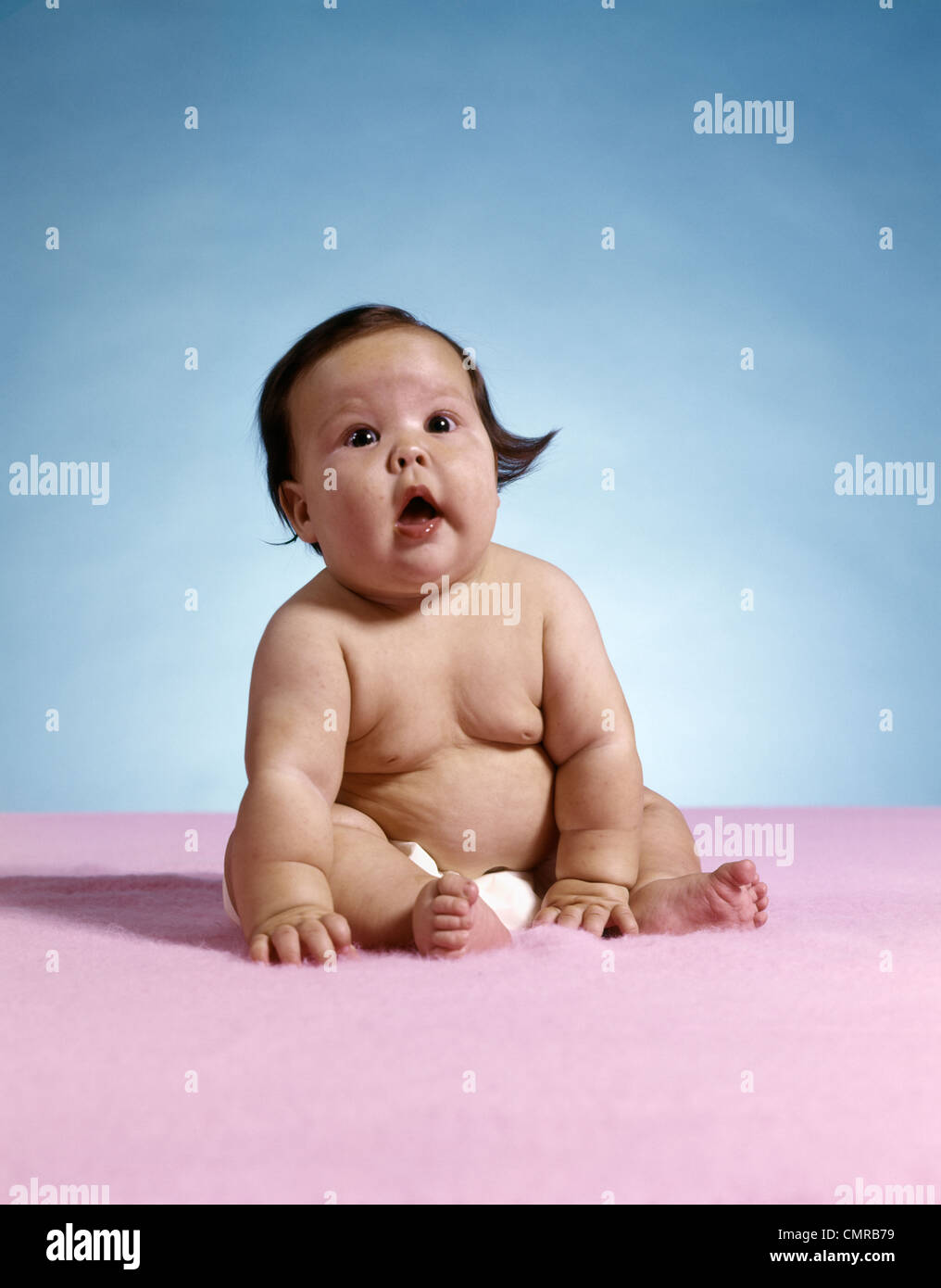 1970s CHUBBY FAT PLUMP BABY OPEN MOUTH WEARING DIAPER SITTING ON PINK RUG Stock Photo