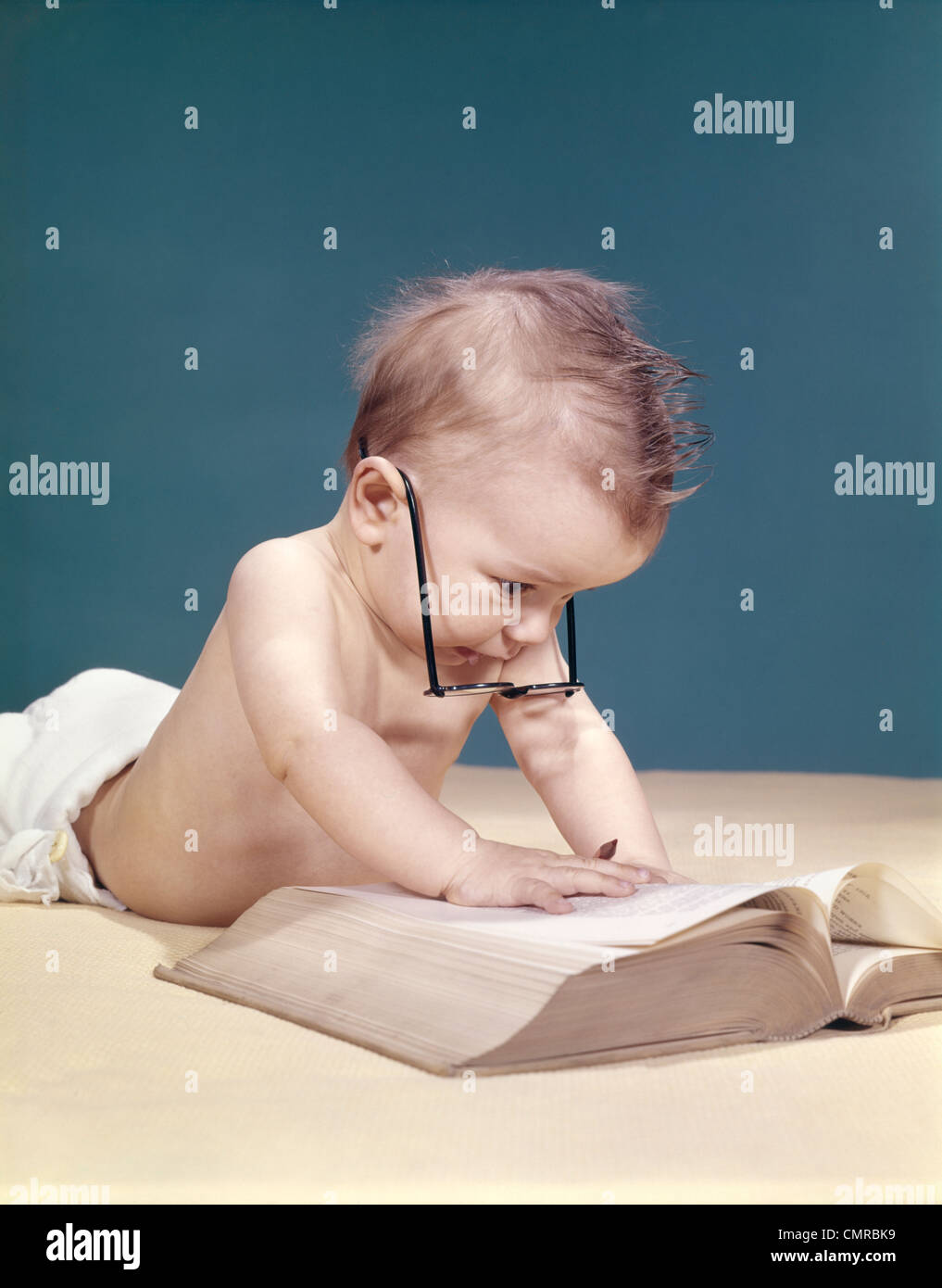 1960s FUNNY BABY WEARING OVERSIZED GLASSES READING BIG BOOK Stock Photo