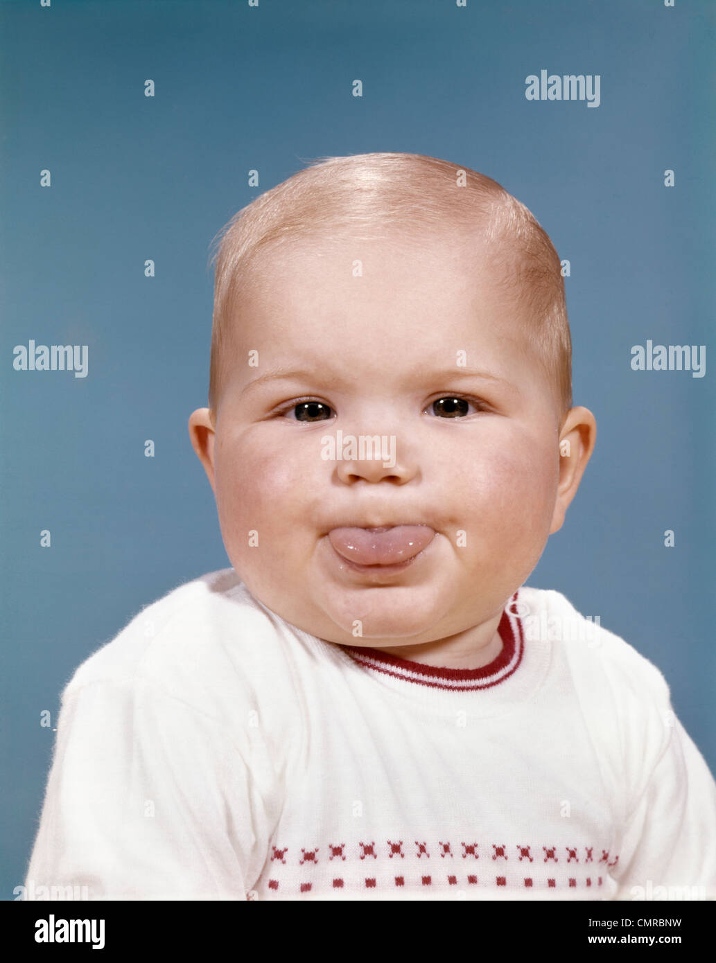 1960s PORTRAIT OF CUTE BLOND CHUBBY BABY LOOKING AT CAMERA STICKING OUT