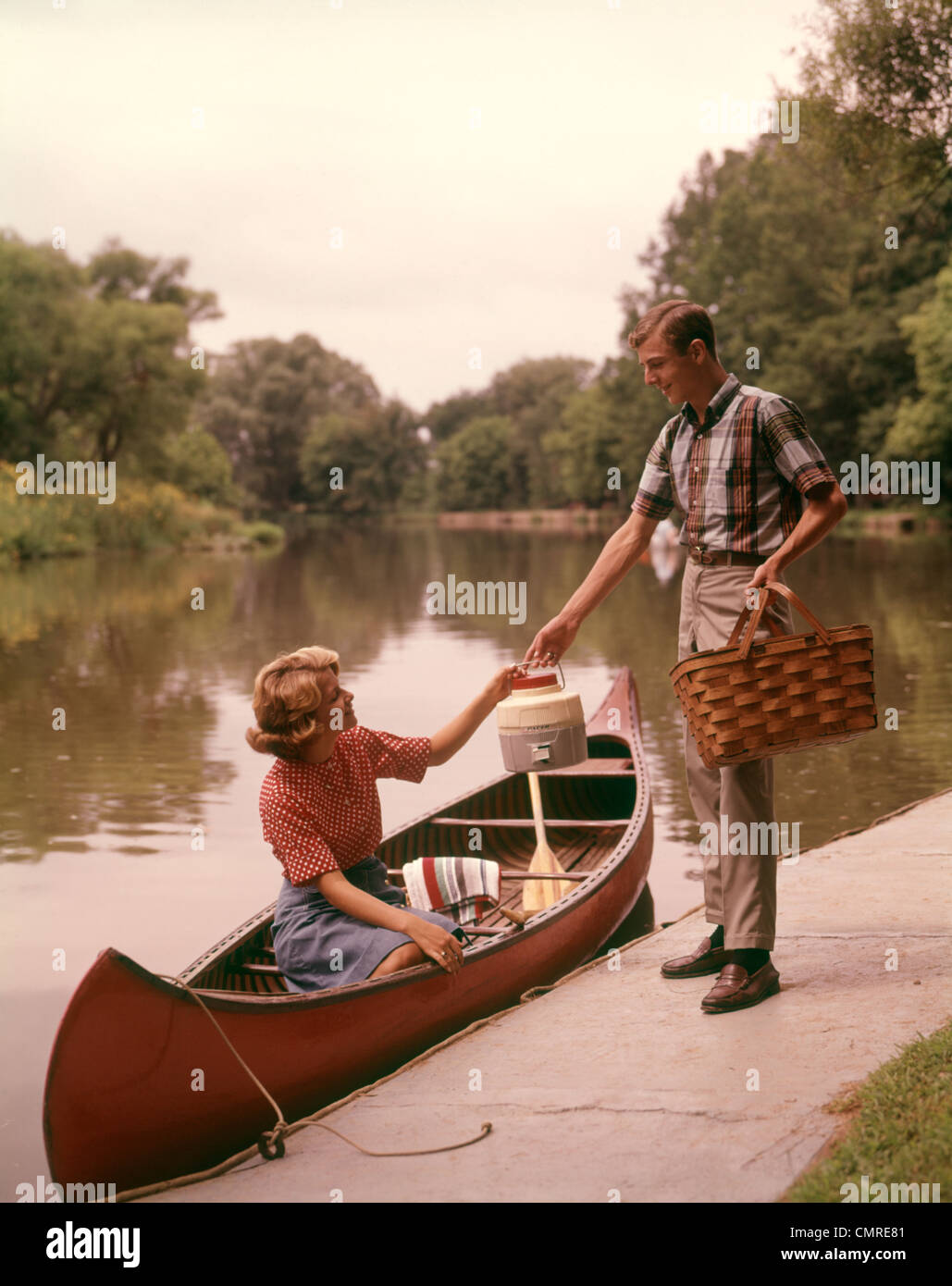 1960s YOUNG COUPLE LOADING PICNIC BASKET THERMOS INTO CANOE Stock Photo
