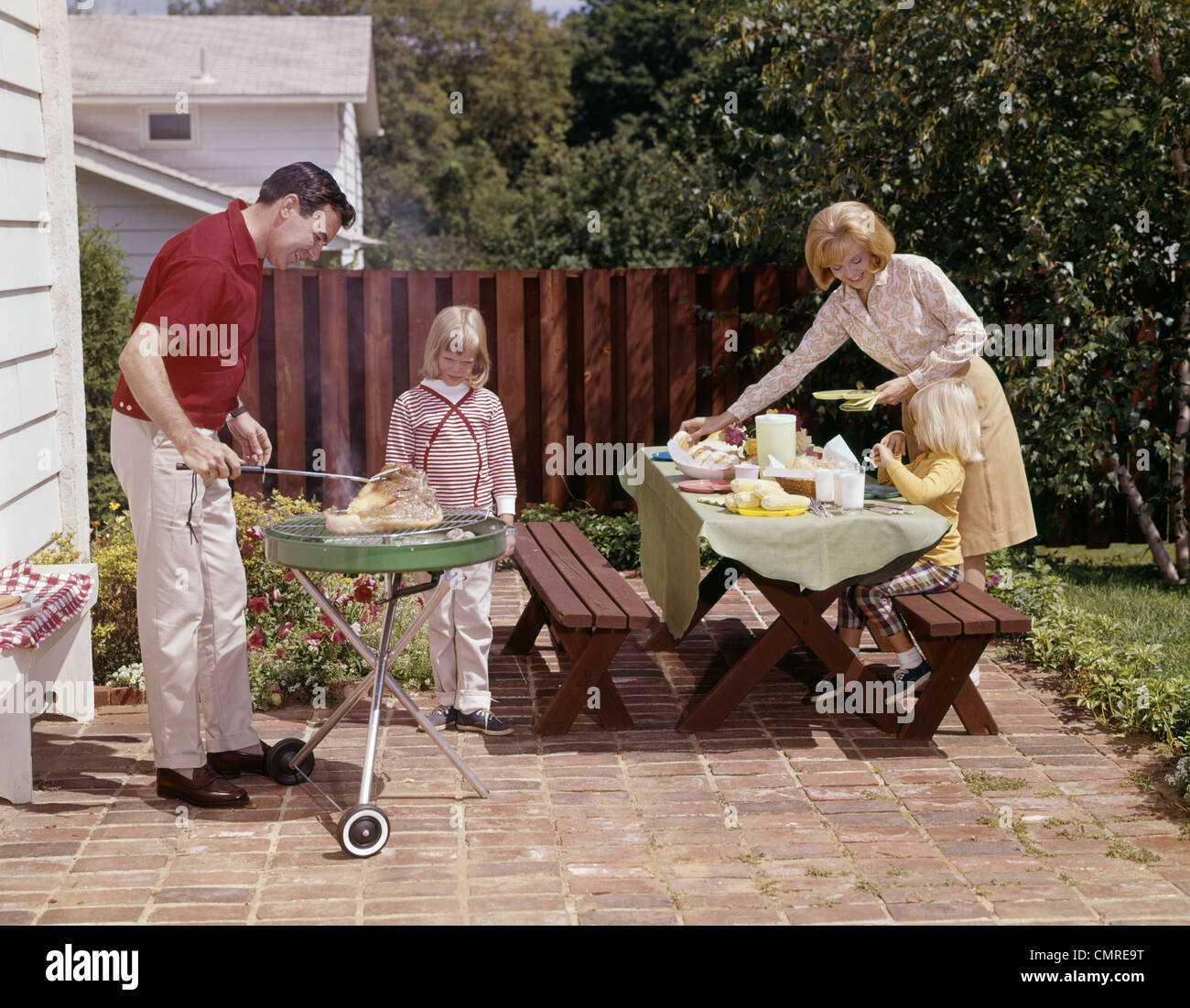 1960s FAMILY FATHER MOTHER TWO DAUGHTERS BARBEQUE IN BACKYARD ON BRICK PATIO Stock Photo