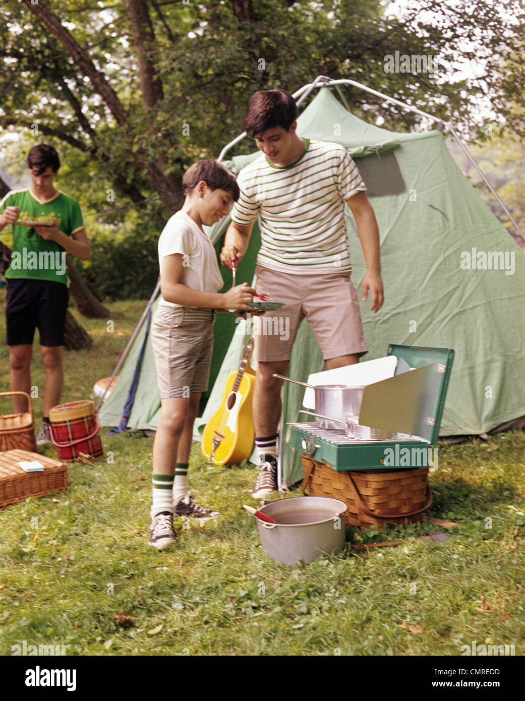 1960s THREE BOYS AT CAMPSITE COOKING CAMP STOVE EATING FOOD TENT TEENS Stock Photo