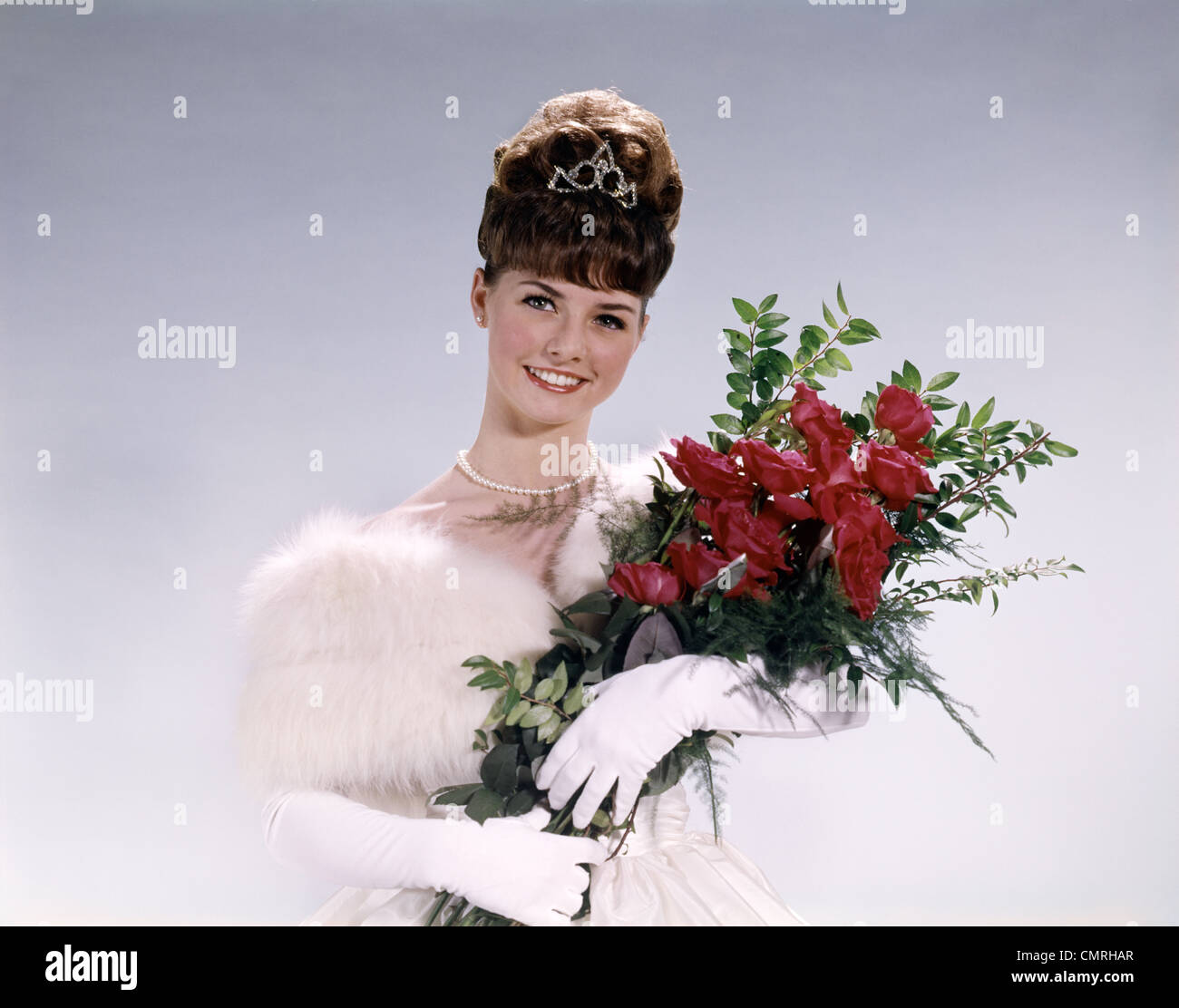 1960s WOMAN PROM QUEEN WEARING WHITE EVENING DRESS HOLDING BOUQUET OF FLOWERS RED ROSES LOOKING AT CAMERA Stock Photo