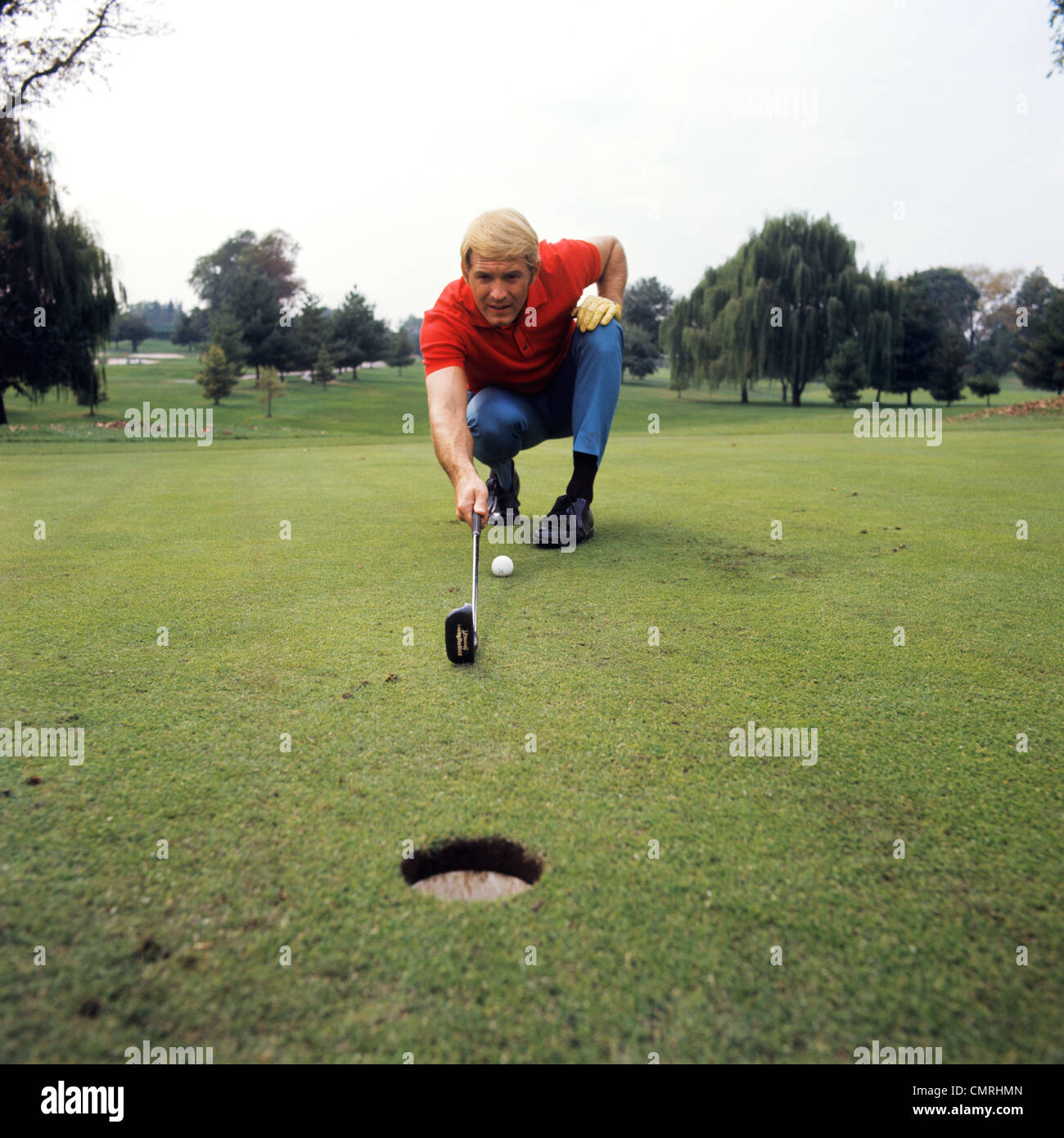 1970s BLOND MAN PLAYING GOLF CROUCHING LINING UP SHOT TO HOLE ON PUTTING GREEN Stock Photo