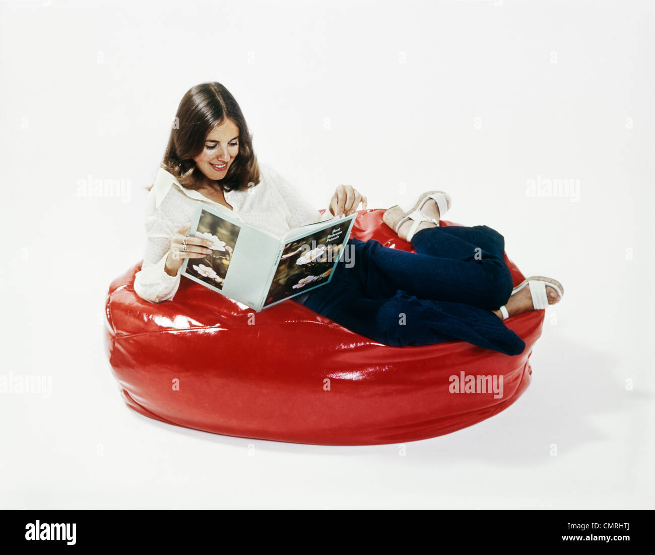 1960s 1970s WOMAN SMILING READING BOOK RECLINING IN A RED BEANBAG CHAIR Stock Photo