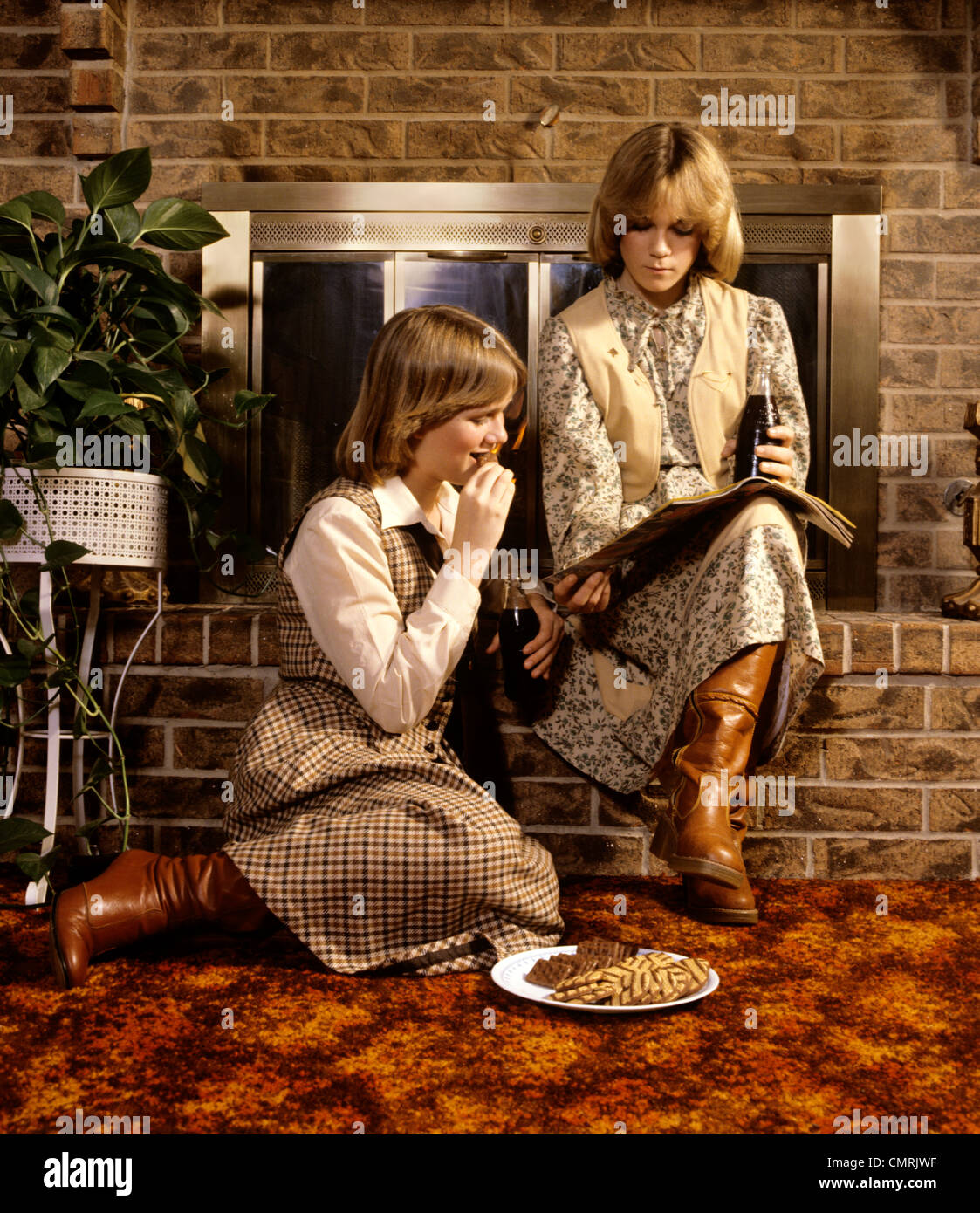 1980 1980s 2 TEENAGE GIRLS SISTERS SIT ON HEARTH READING MAGAZINE EAT  COOKIES FASHION CLOTHES TEENS BOOTS SKIRTS RETRO Stock Photo - Alamy