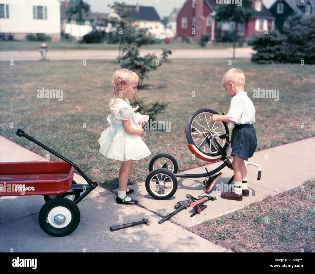 LITTLE GIRL HOLDING DOLL WATCHING LITTLE BOY REPAIR BICYCLE WITH TOOL OUTDOOR Stock Photo