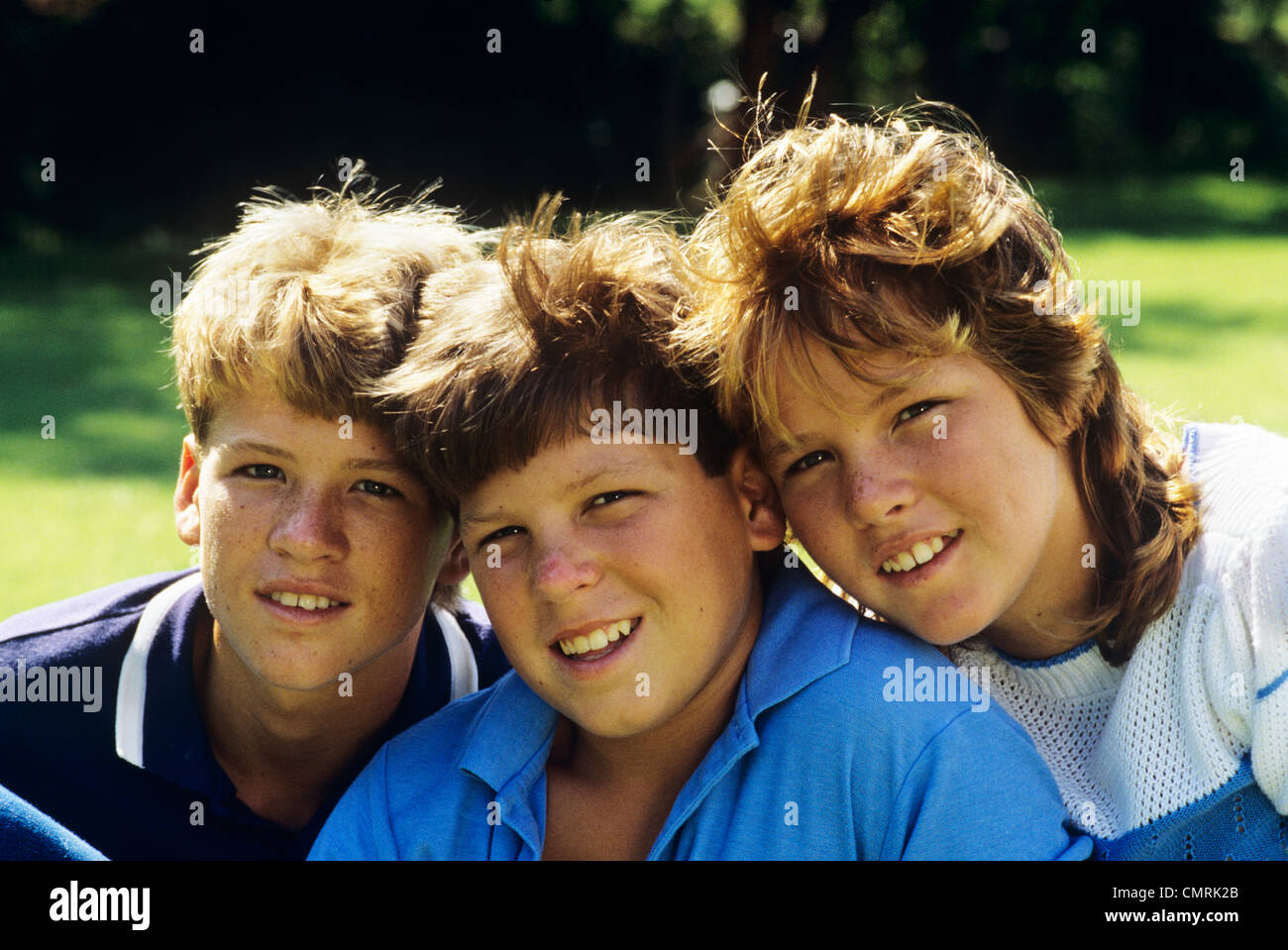 1980s TWO BROTHERS AND THEIR SISTER POSED HEADS TOGETHER SMILING LOOKING AT CAMERA Stock Photo