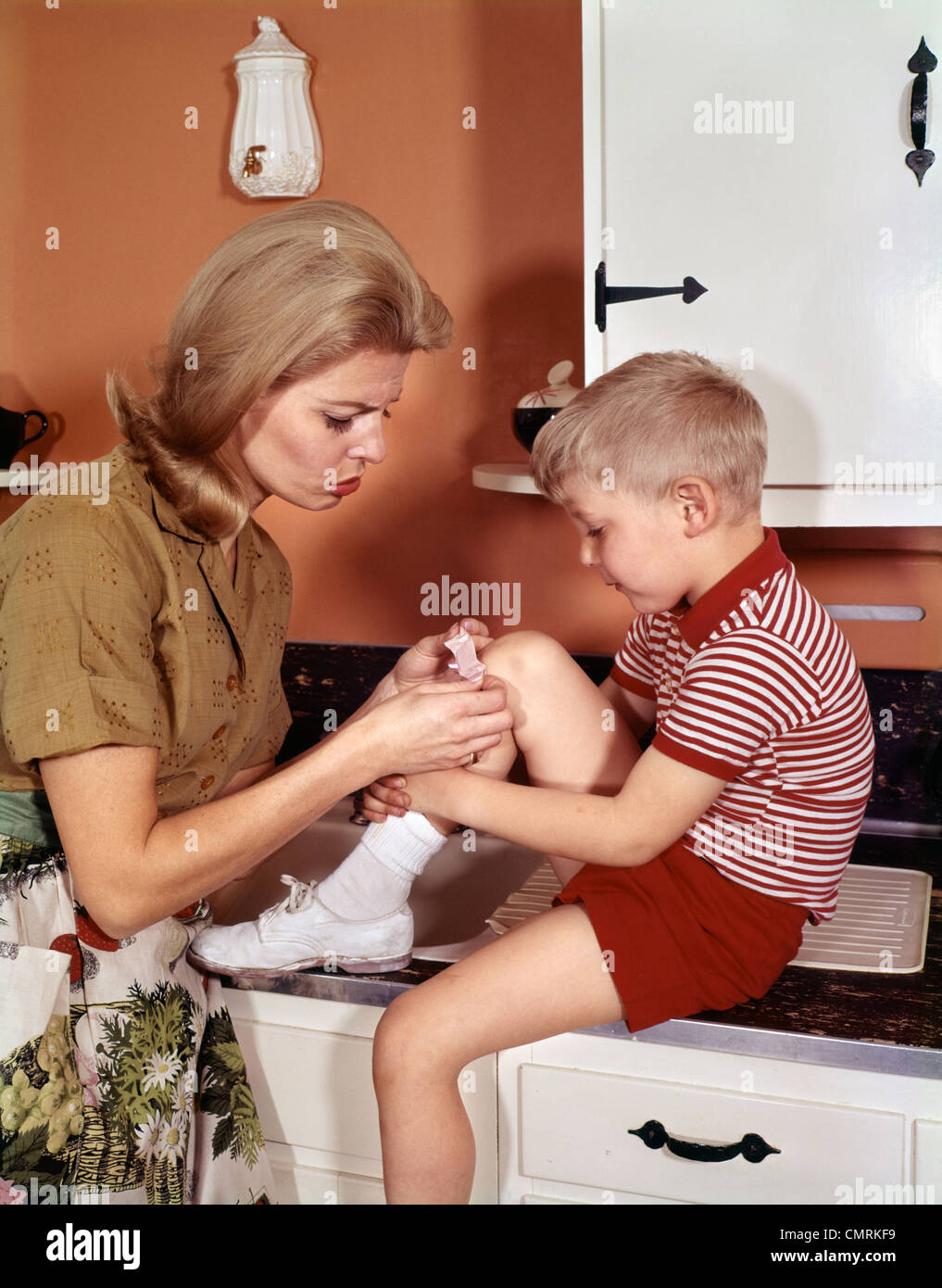 MOTHER GIVING FIRST-AID PUTTING BAND-AID ON KNEE OF SON IN KITCHEN 1960s Stock Photo