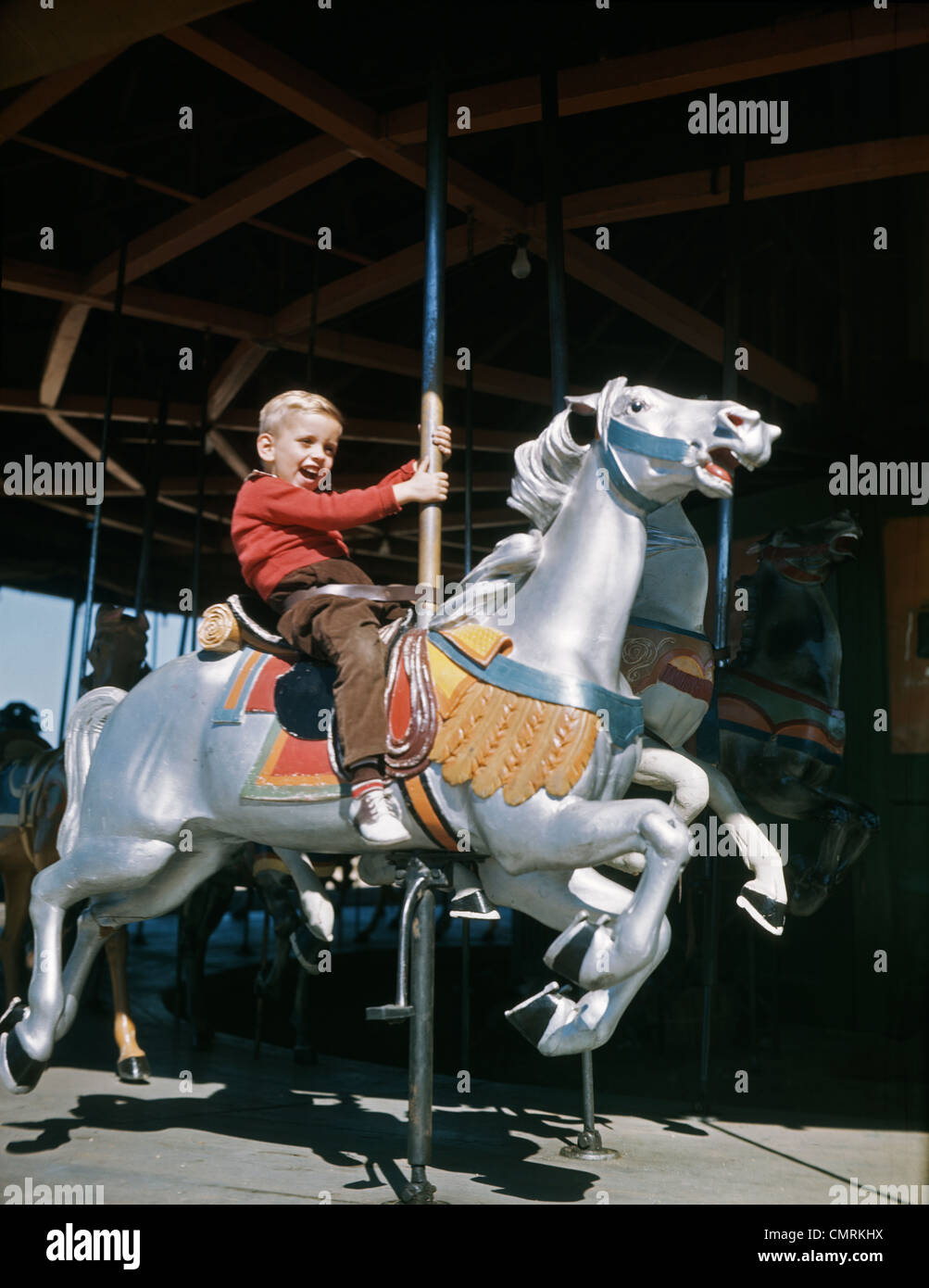 1950s LITTLE EXCITED BOY RIDING CARVED WOOD CAROUSEL MERRY-GO-ROUND HORSE AT AN AMUSEMENT PARK CARNIVAL Stock Photo