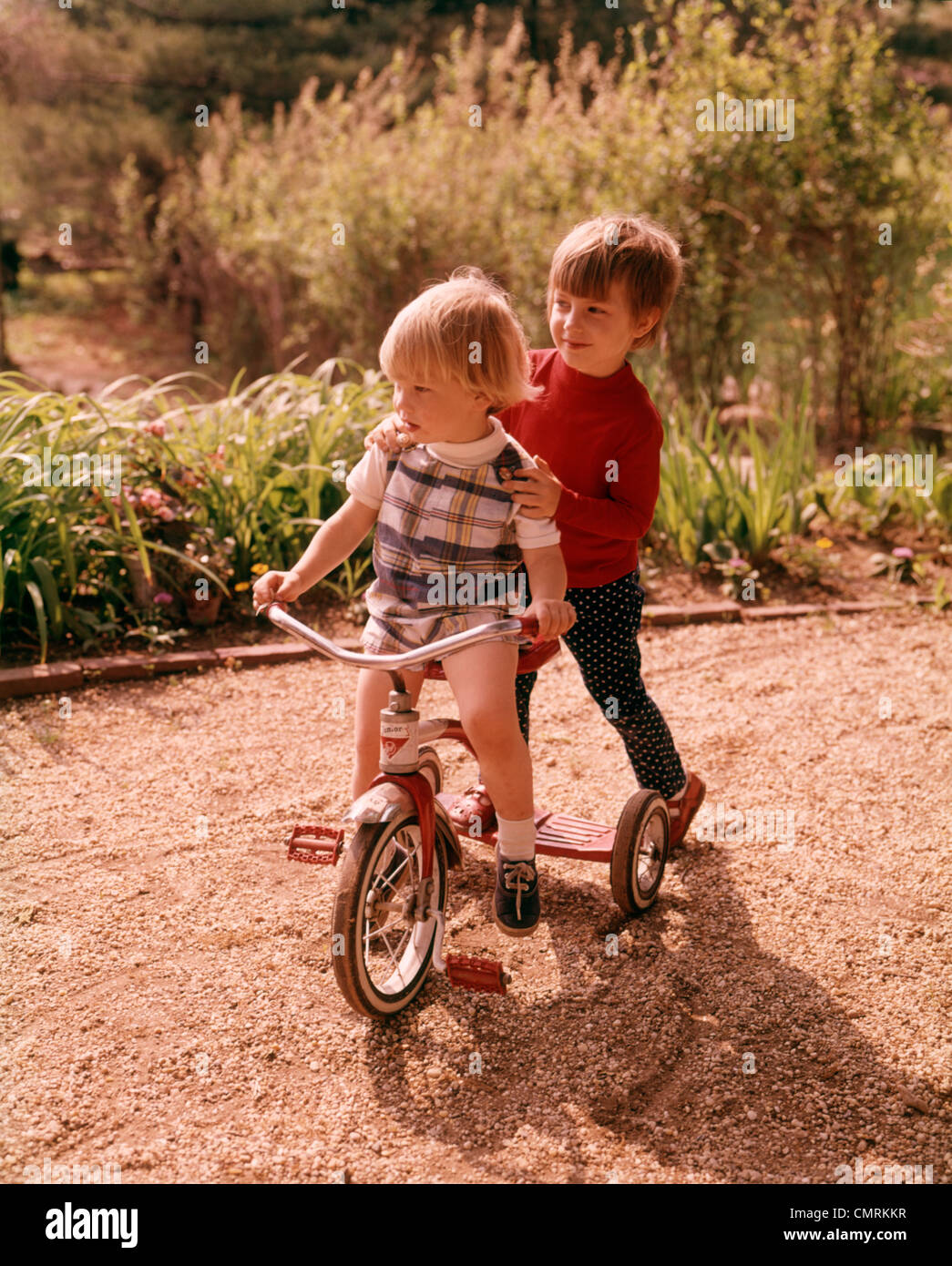 1970 1970 TWO KIDS RIDING TRICYCLE BIKE BICYCLE PLAYING PLAY TODDLER BOY GIRL PLAY RETRO Stock Photo