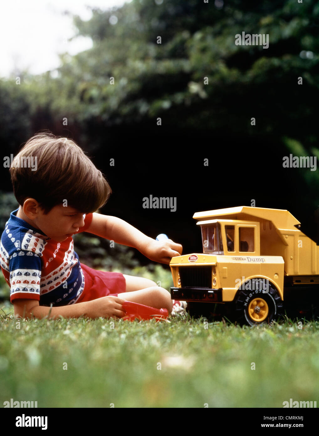 1970s BOY IN GRASS PLAYING WITH TONKA TOY YELLOW DUMP TRUCK Stock Photo