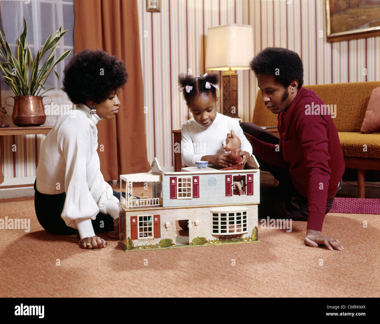 1970s AFRICAN-AMERICAN FAMILY MOTHER FATHER GIRL DAUGHTER PLAING ON FLOOR WITH DOLLHOUSE DOLL HOUSE RETRO Stock Photo