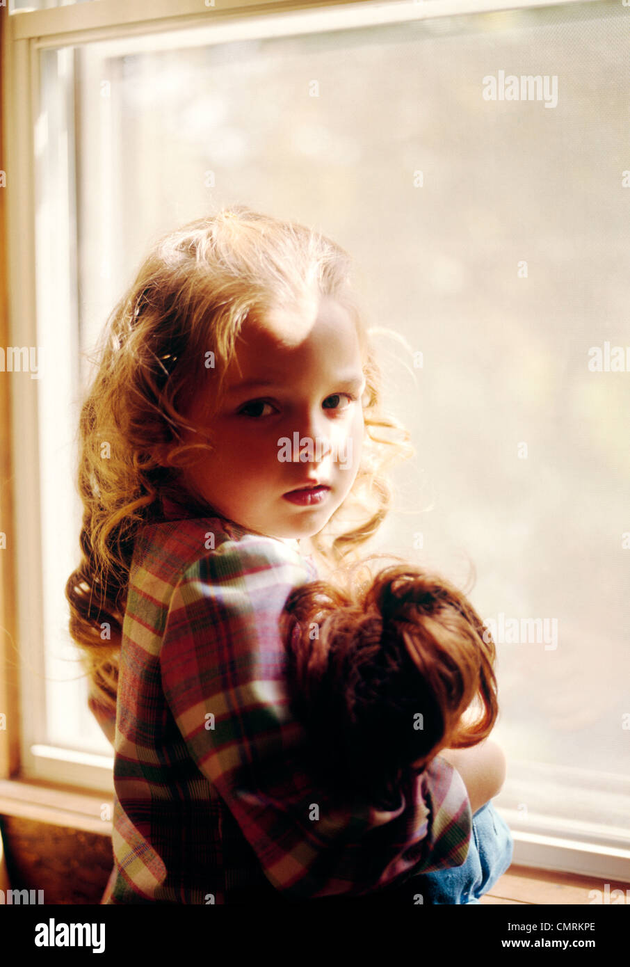 1970 1970s RETRO BLONDE GIRL HOLDING DOLL STANDING BY WINDOW SAD LONELY MELANCHOLY Stock Photo