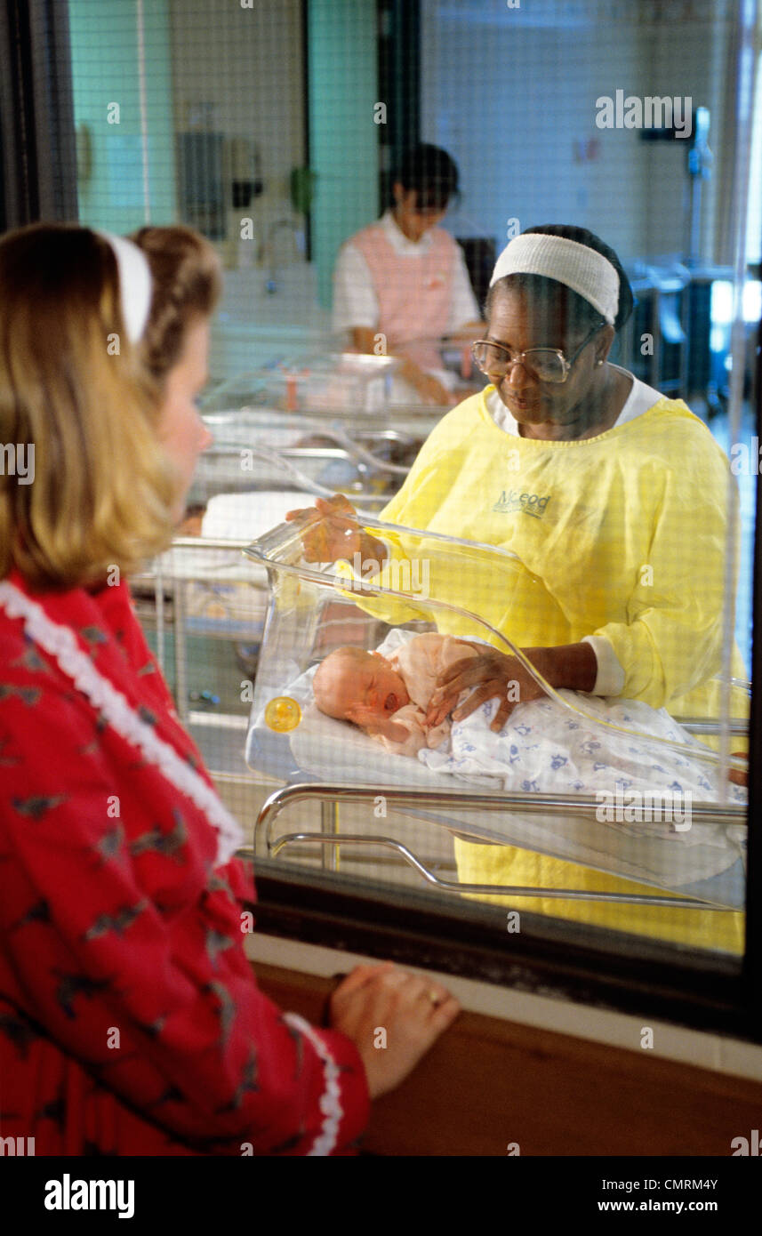 NURSE IN HOSPITAL NURSERY SHOWING BABY TO MOTHER THROUGH WINDOW Stock Photo