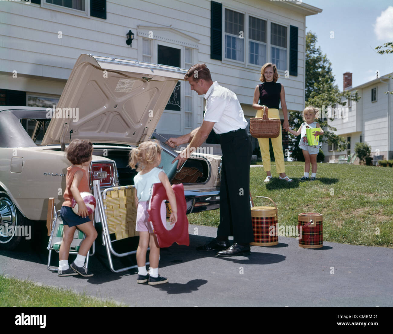 FAMILY MOTHER FATHER THREE DAUGHTERS PACKING LUGGAGE INTO CAR FOR VACATION OUTDOOR 1970s Stock Photo
