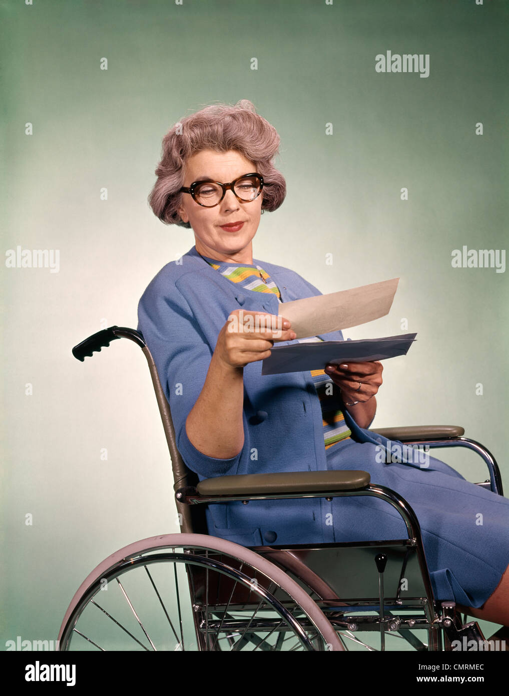 1960s SENIOR WOMAN WEARING GLASSES SITTING IN WHEELCHAIR READING LETTER INSURANCE CHECK HEALTHCARE PATIENT Stock Photo