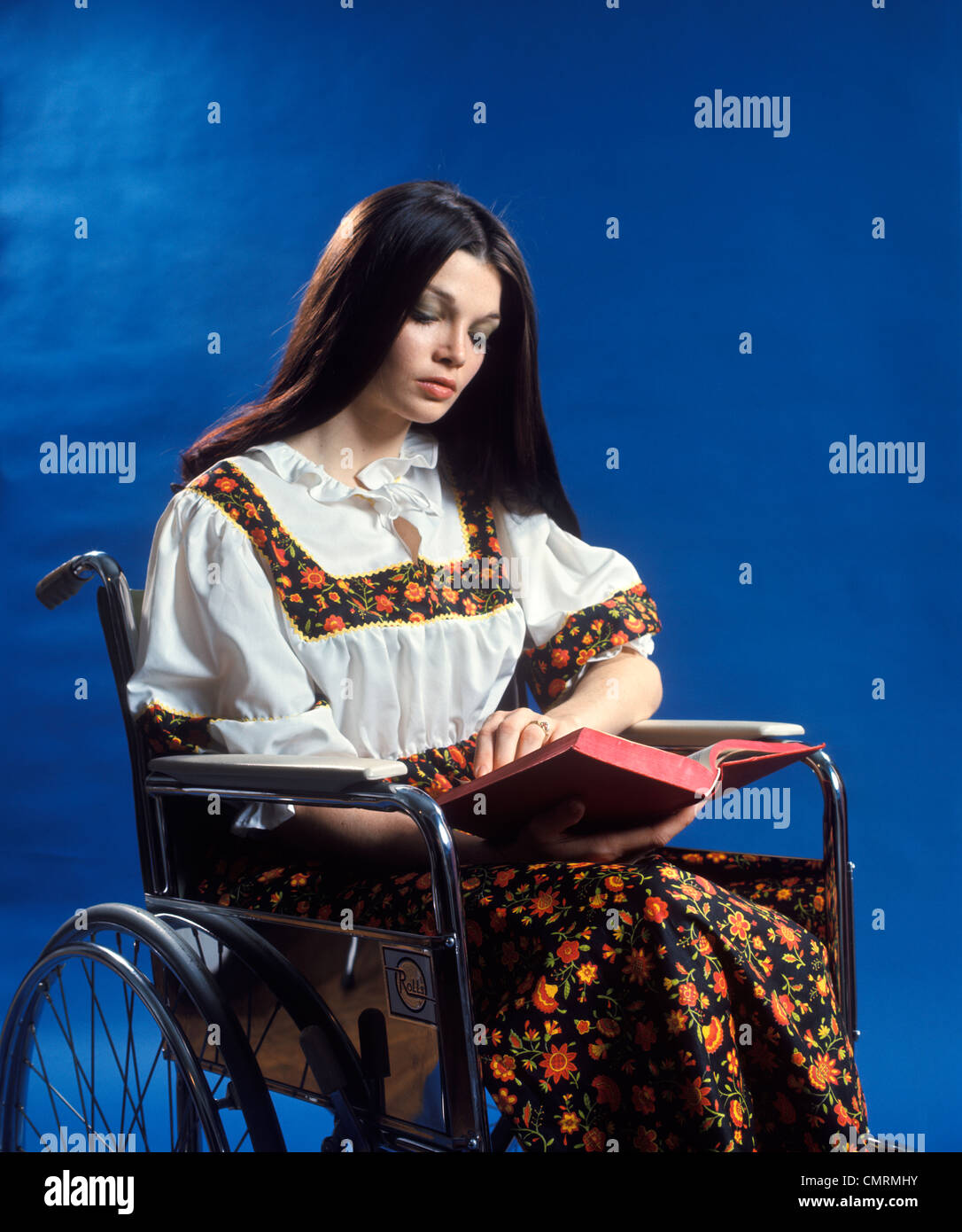 1970 1970s RETRO WOMAN LONG BROWN HAIR SITTING IN WHEELCHAIR READING BOOK PATIENT HEALTHCARE Stock Photo