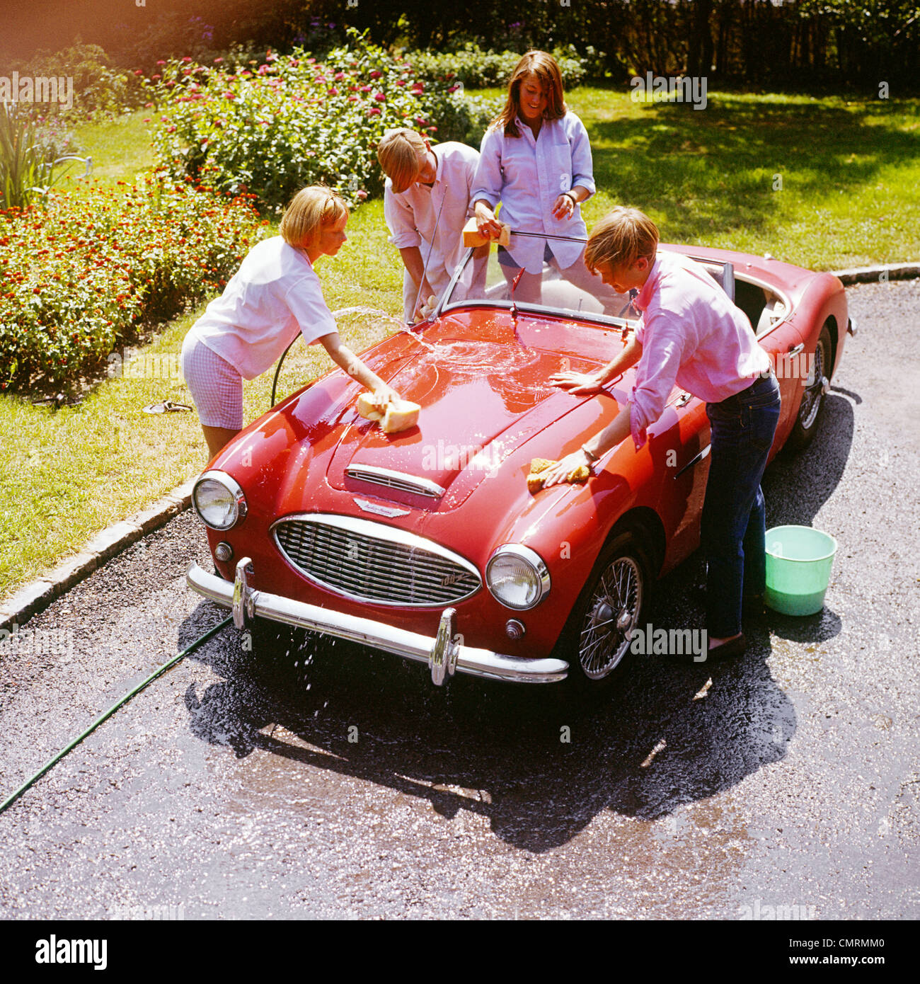 1970s FOUR TEENAGERS WASHING RED AUSTIN HEALEY SPORTS CONVERTIBLE AUTOMOBILE MAN WOMAN OVERHEAD OUTDOOR Stock Photo