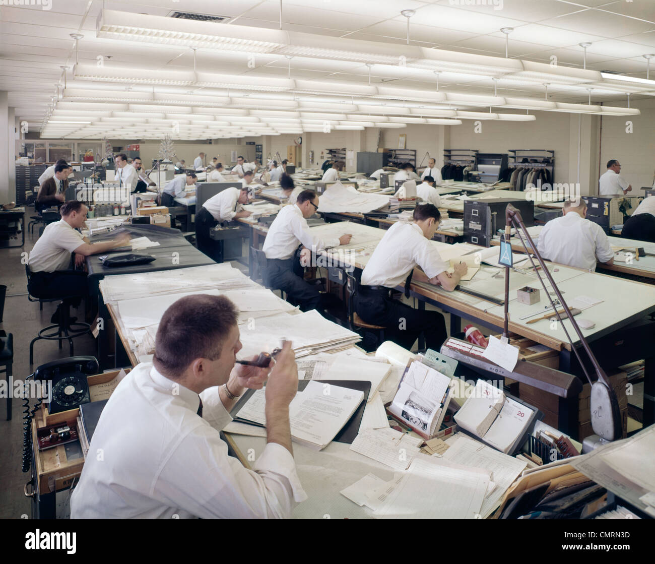 1960s OFFICE FULL OF DRAFTING TABLES DESIGNER MAN LIGHTING-UP A PIPE OF TOBACCO INDOOR INDUSTRY Stock Photo