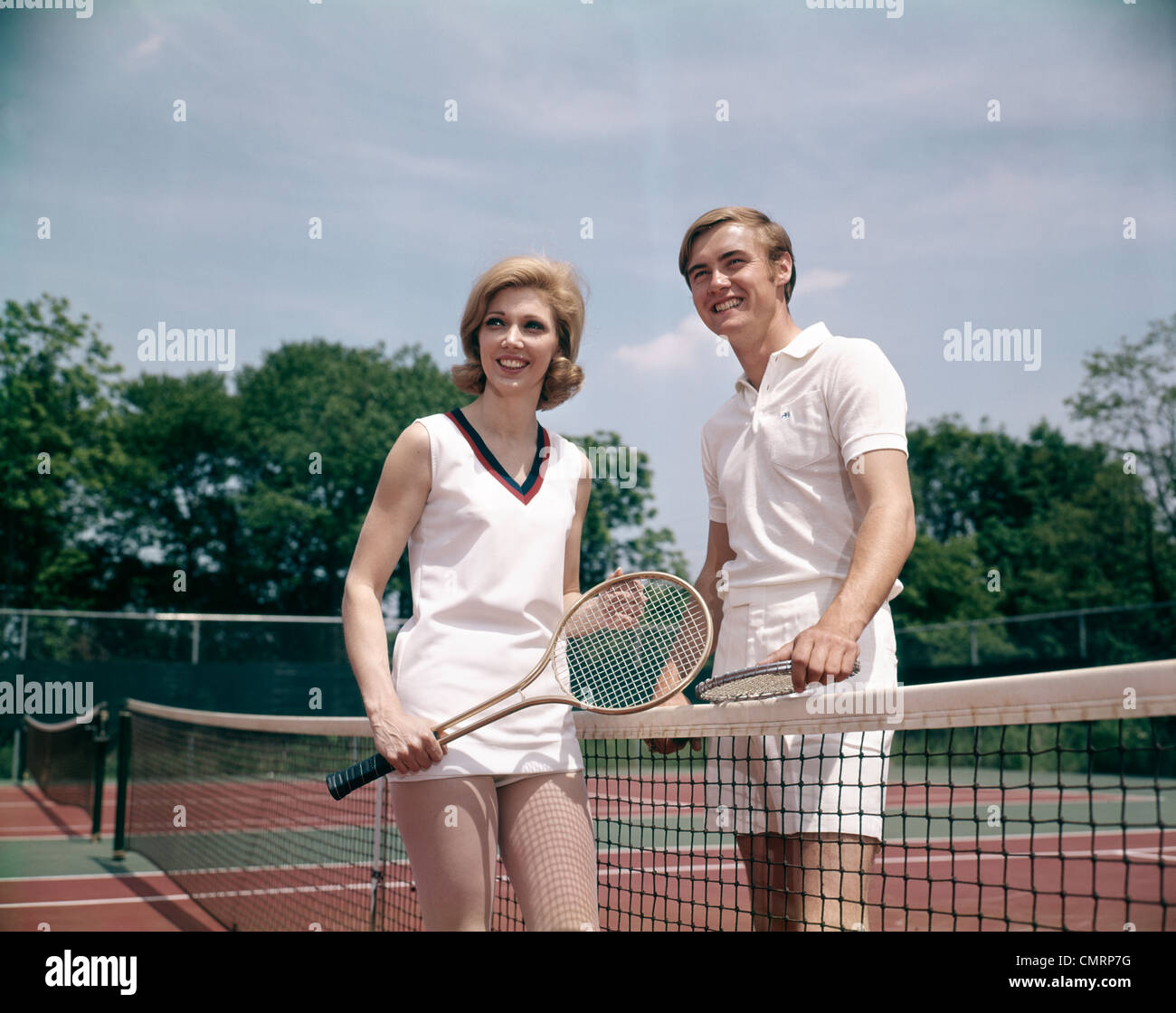 1970 1970s RETRO SMILING COUPLE MAN WOMAN STANDING ON OPPOSITE SIDES OF NET TENNIS COURT HOLDING RACKETS RECREATION Stock Photo