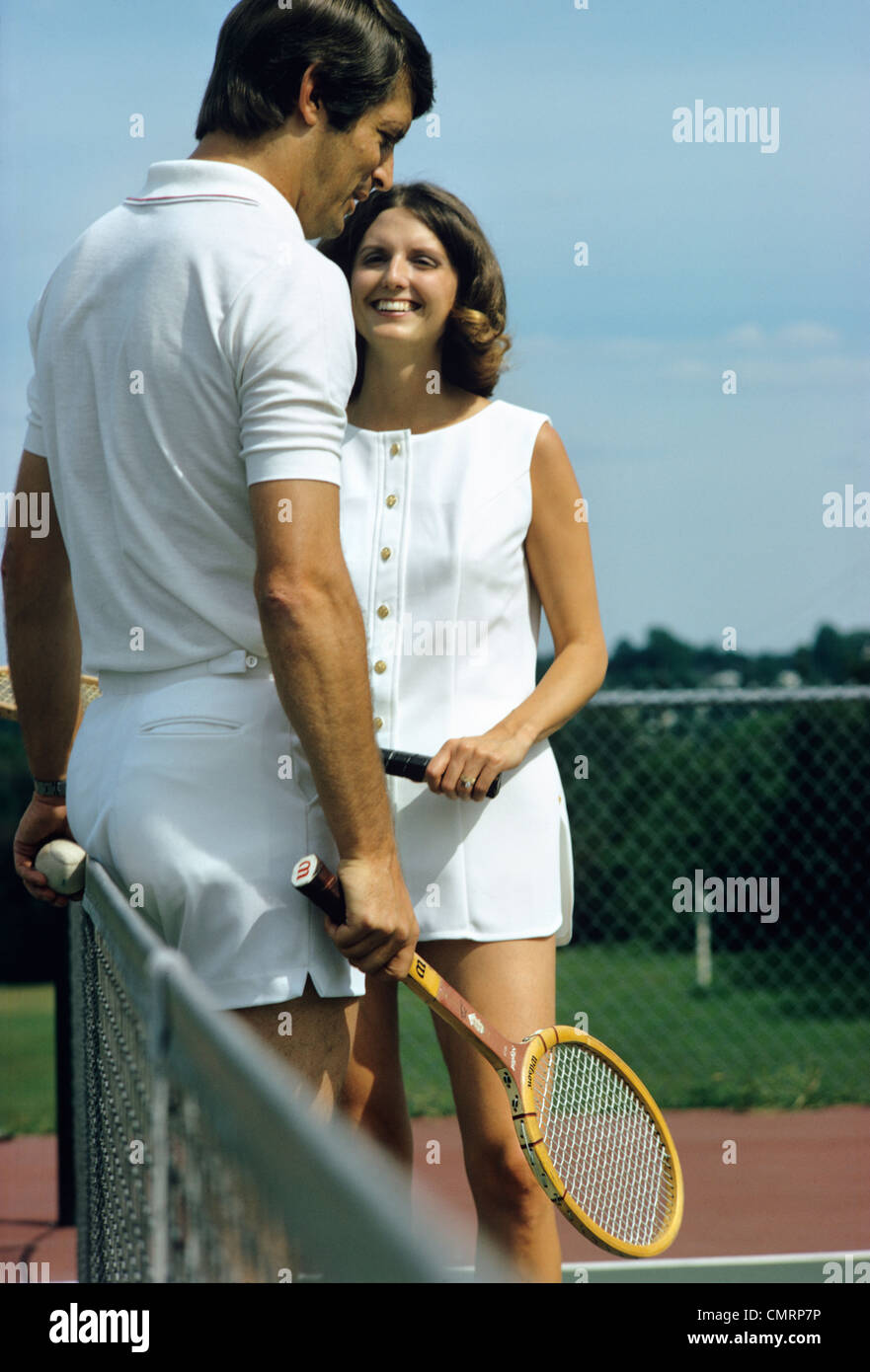 1970 1970s RETRO SMILING COUPLE MAN WOMAN STANDING BY NET ON TENNIS COURT HOLDING RACKETS RECREATION EXERCISE Stock Photo