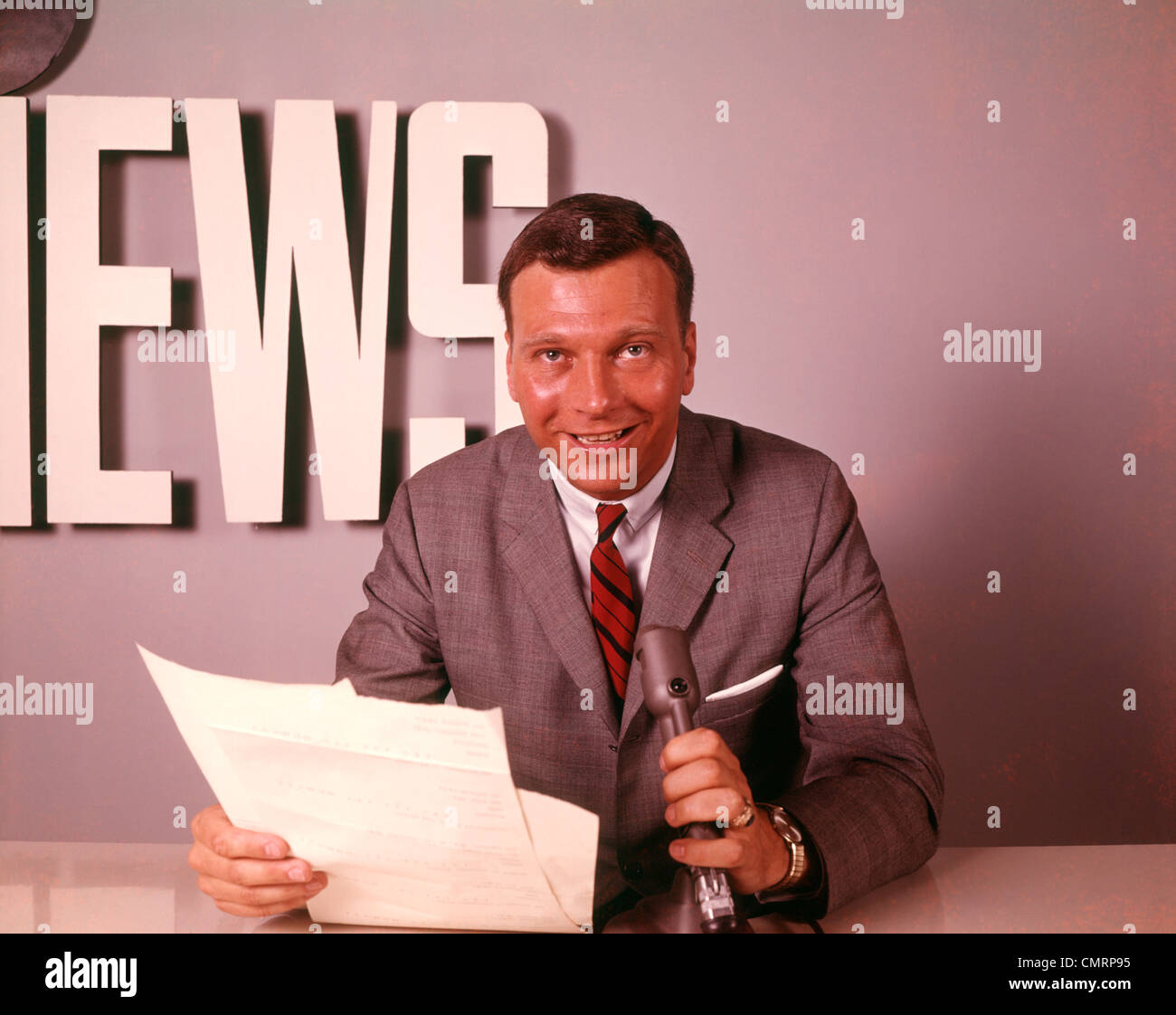 1960 1960s SMILING REPORTER NEWSMAN ANNOUNCER AT NEWS DESK MICROPHONE PAPERS READING REPORT BROADCASTING RETRO Stock Photo