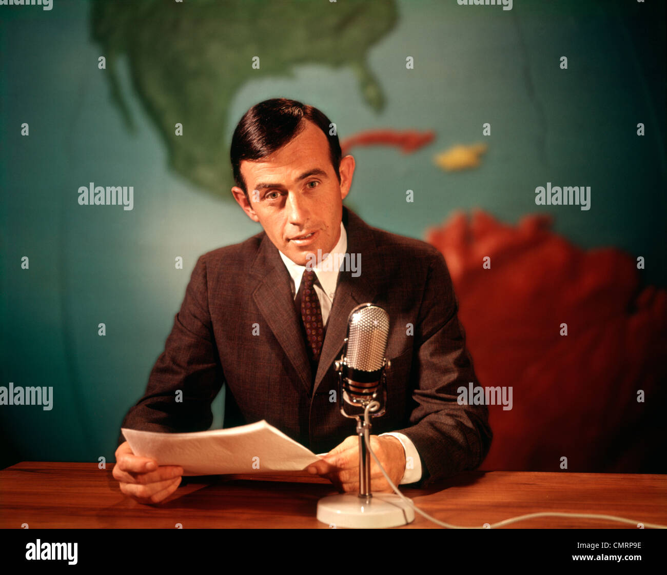 1960 1960s SERIOUS NEWS REPORTER READING NEWS INTO MICROPHONE WITH GLOBAL MAP BACKGROUND REPORT NEWSMAN MAN MEN RETRO Stock Photo