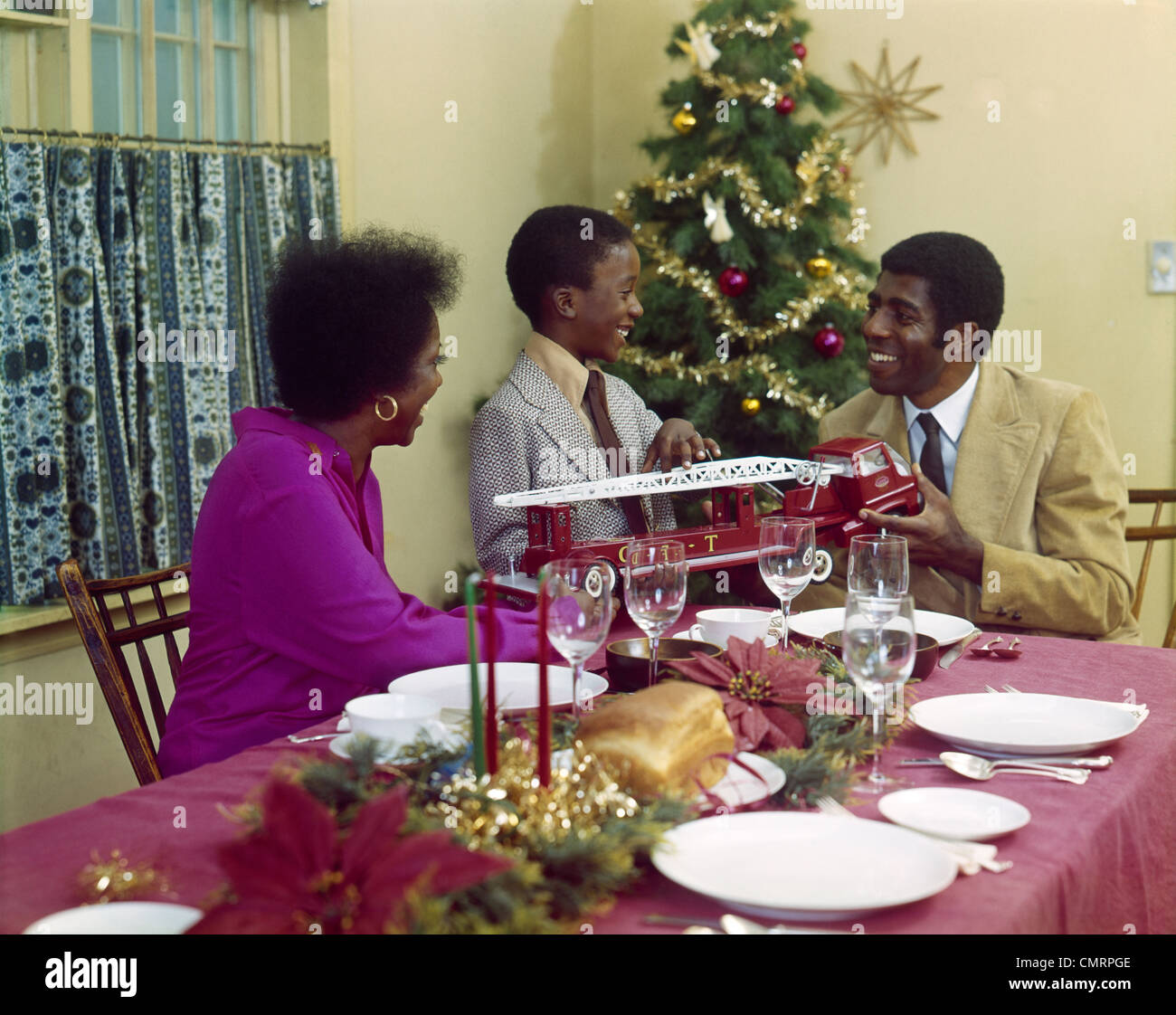 1970s AFRICAN-AMERICAN FAMILY MOTHER FATHER SON DINING TABLE WITH TOY FIRE TRUCK CHRISTMAS TREE Stock Photo