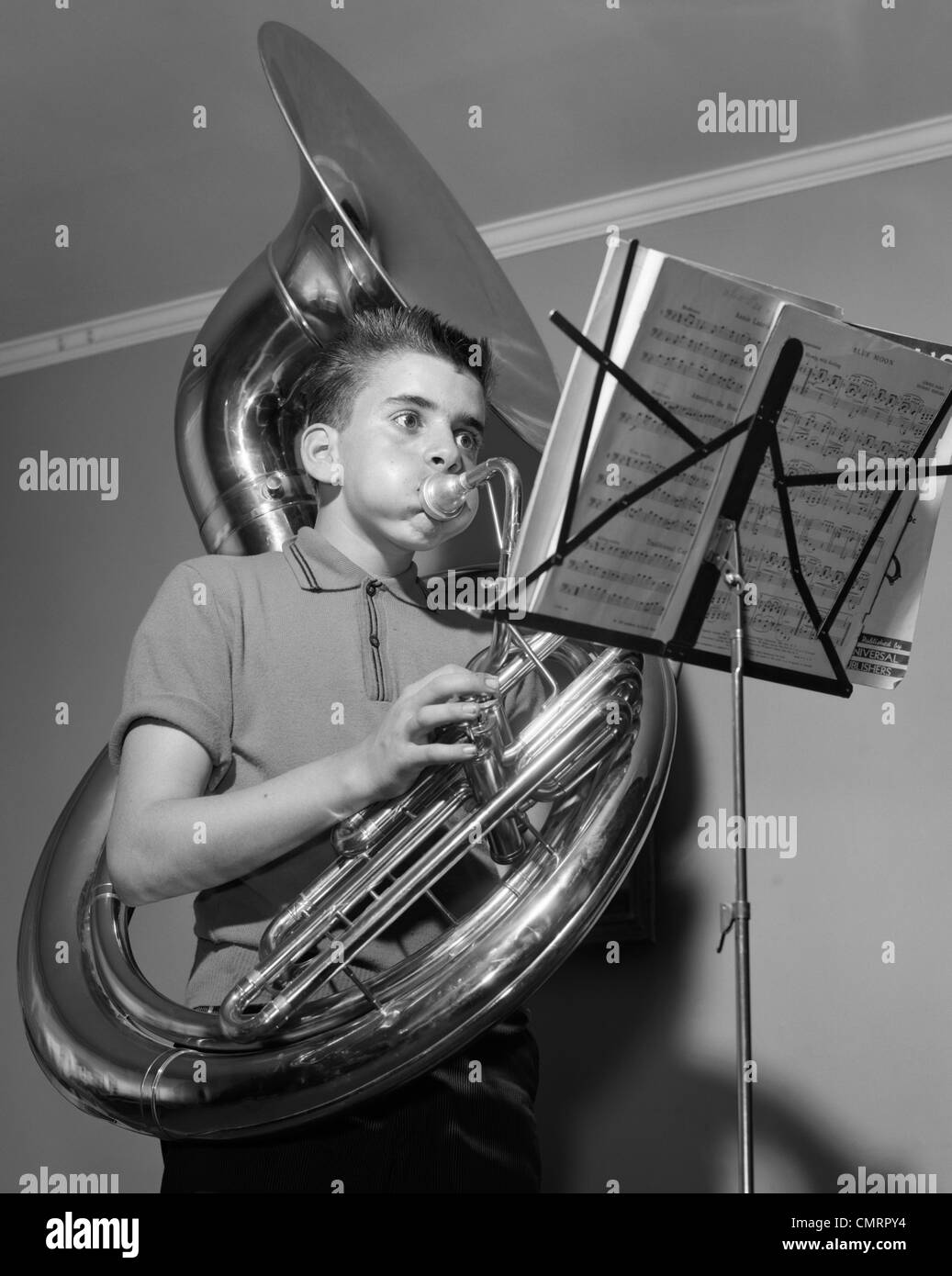 1960s BOY PLAYING THE TUBA WHILE READING FROM SHEET MUSIC ON A MUSIC STAND INSIDE Stock Photo