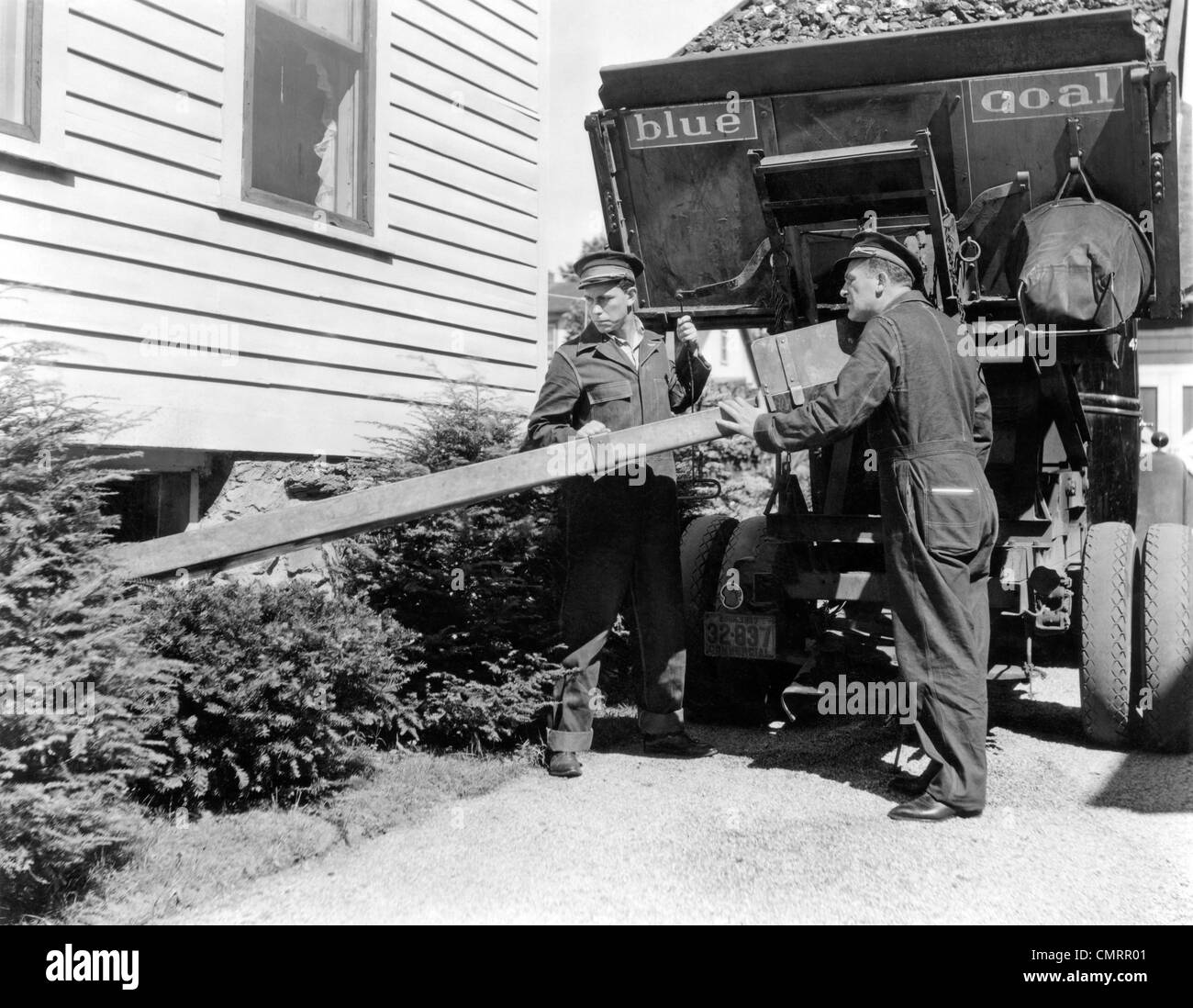 1930s 1940s COAL TRUCK WITH TWO SERVICE MEN MAKING HOME DELIVERY COAL CHUTE INTO BASEMENT Stock Photo