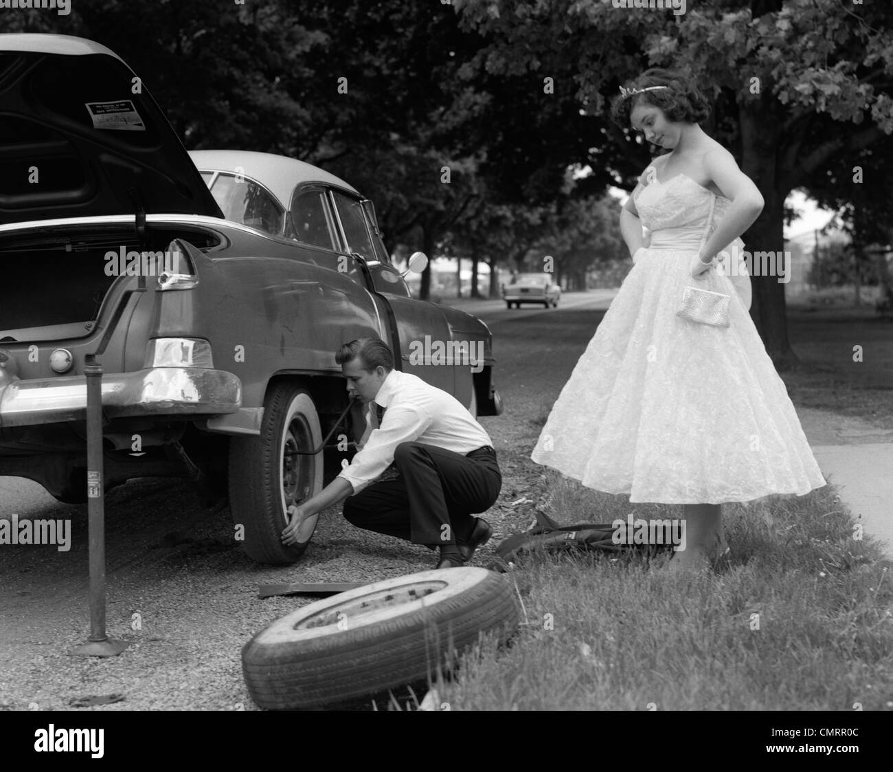 1950s TEENAGE BOY CROUCHING DOWN CHANGING FLAT TIRE PROM DATE WATCHING WITH HANDS ON HIPS Stock Photo