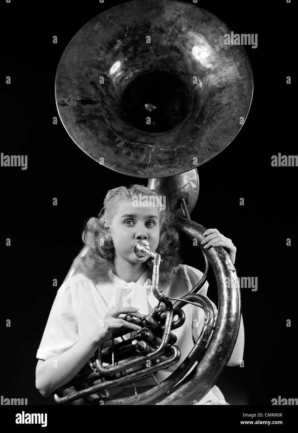 1940s TEENAGE WOMAN BLOWING AND PLAYING TUBA HORN MUSICAL INSTRUMENT WITH EYES WIDE OPEN LOOKING AT CAMERA Stock Photo