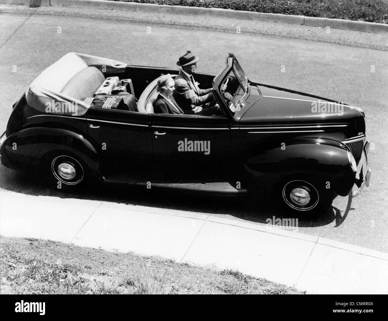 1930s 1940s COUPLE HUSBAND AND WIFE DRIVING 1938 CONVERTIBLE FOUR DOOR SEDAN AUTOMOBILE WITH LUGGAGE IN BACK SEAT Stock Photo