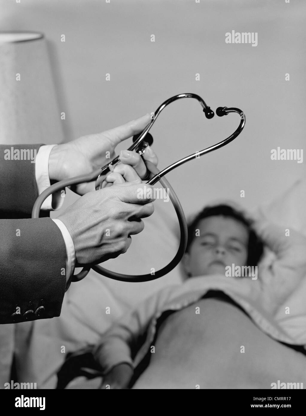 1960s DOCTOR HANDS HOLDING STETHOSCOPE LITTLE GIRL PATIENT SLEEPING AT HOME Stock Photo