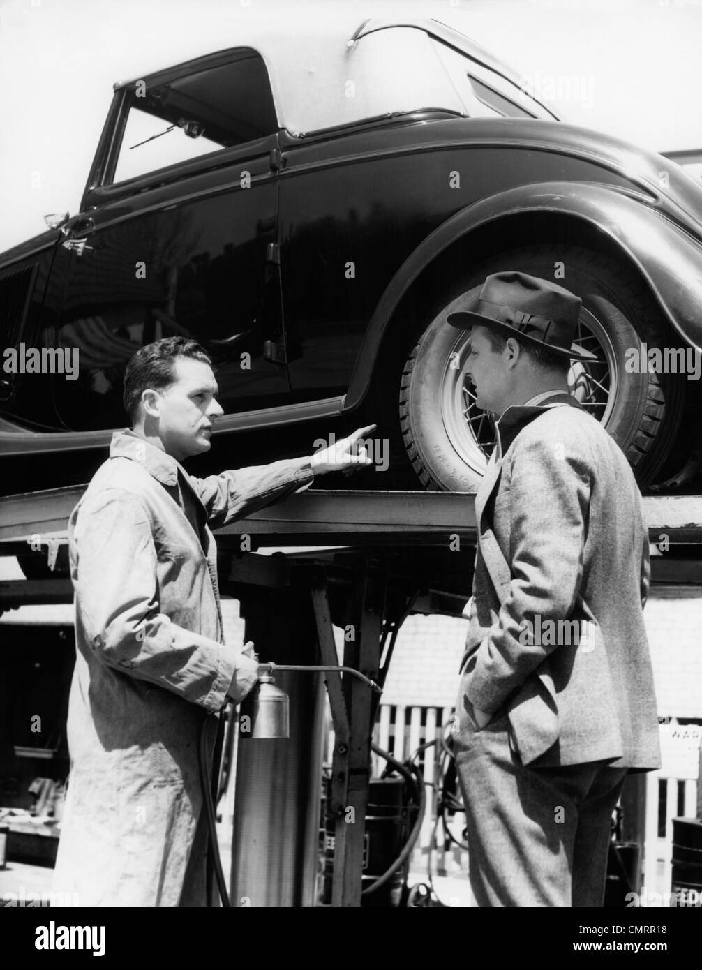 1930s SERVICE STATION ATTENDANT IN COVERALLS HOLDING OIL CAN POINTING AT PROBLEM CONVERTIBLE ON LIFT TALKING TO OWNER Stock Photo