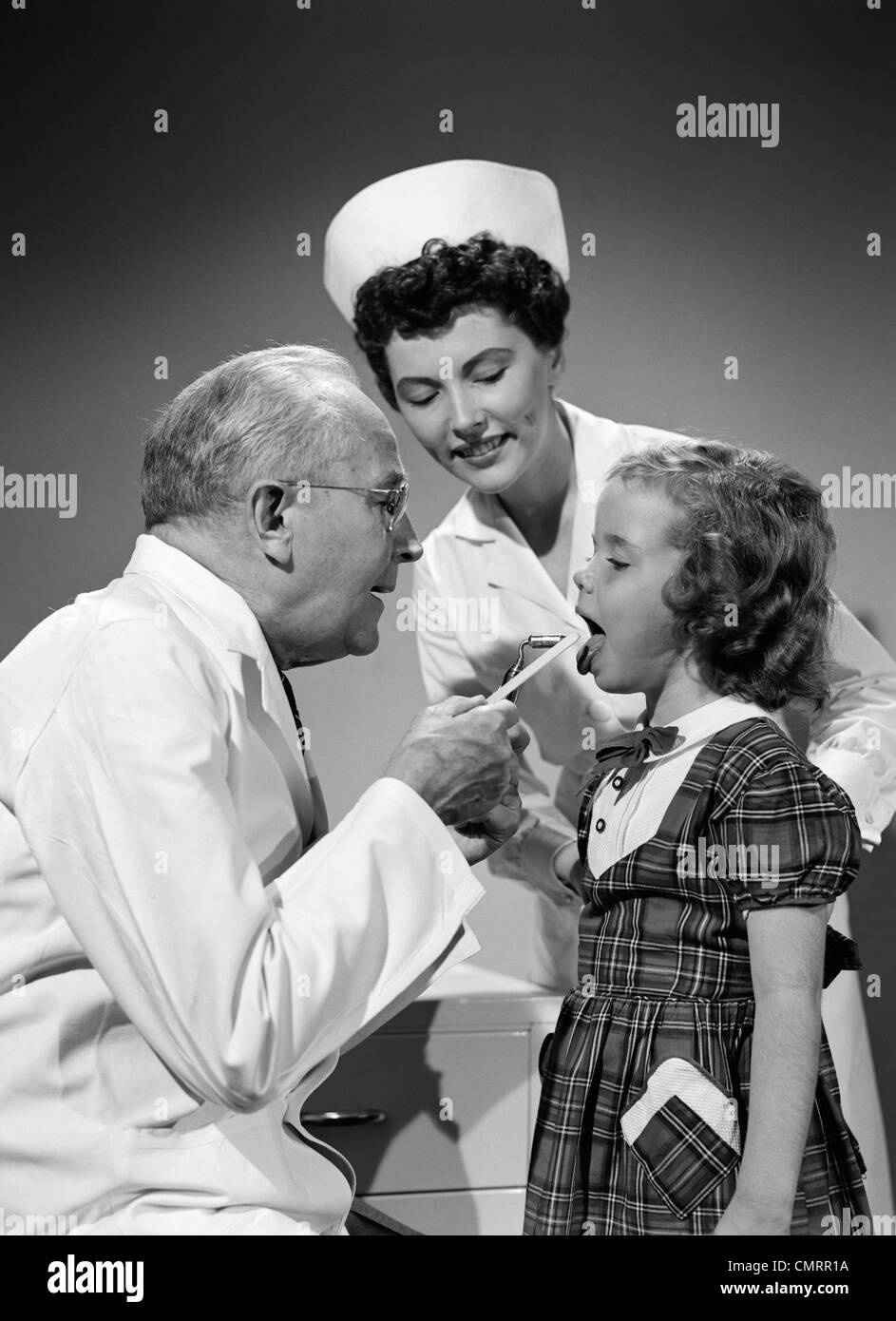 1950s DOCTOR EXAMINING MOUTH OF LITTLE GIRL WITH TONGUE DEPRESSOR WHILE NURSE IS LOOKING ON Stock Photo