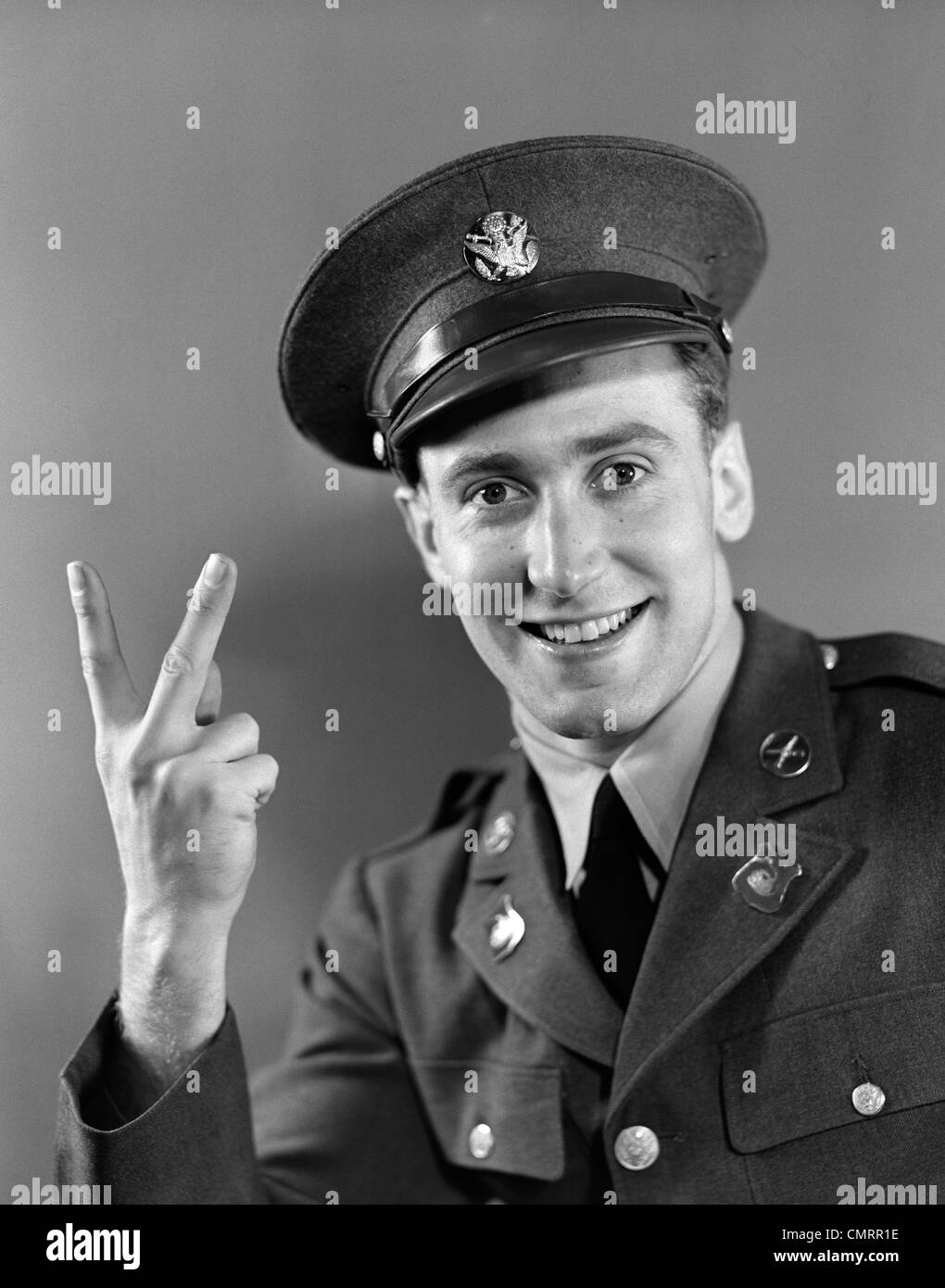 1940s PORTRAIT OF YOUNG ARMY MAN IN UNIFORM SMILING & MAKING VICTORY SIGN WITH HAND Stock Photo