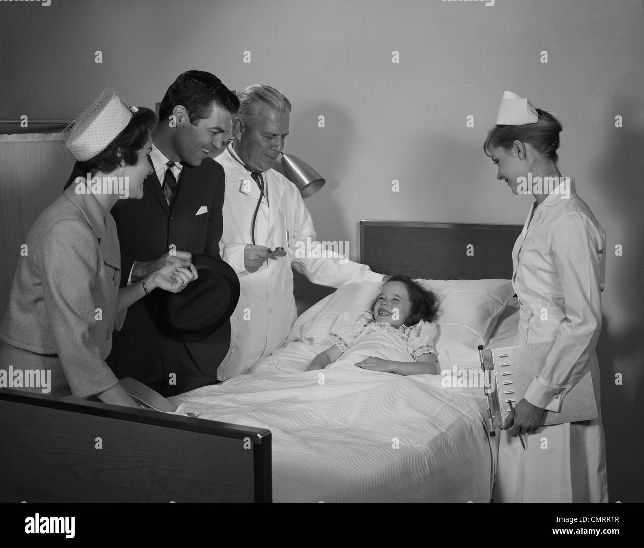 1960s MAN DOCTOR WOMAN NURSE MOTHER FATHER SEEING VISITING SMILING LITTLE GIRL PATIENT IN HOSPITAL BED Stock Photo