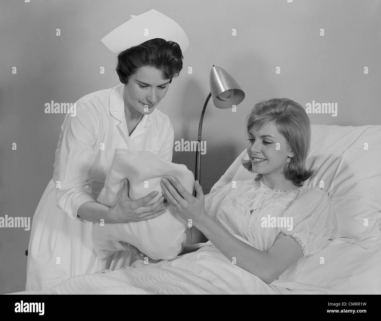 1960s WOMAN NURSE GIVING BABY TO SMILING MOTHER PATIENT SITTING IN HOSPITAL BED Stock Photo