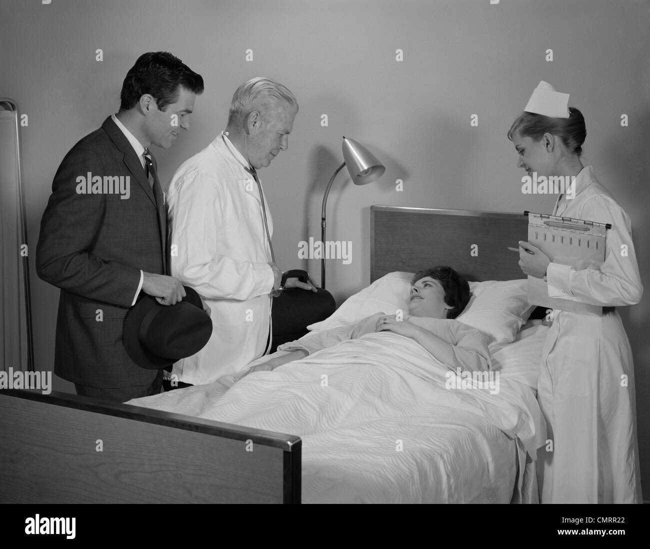 1960s DOCTOR NURSE HUSBAND TALKING WITH FEMALE PATIENT IN HOSPITAL BED Stock Photo