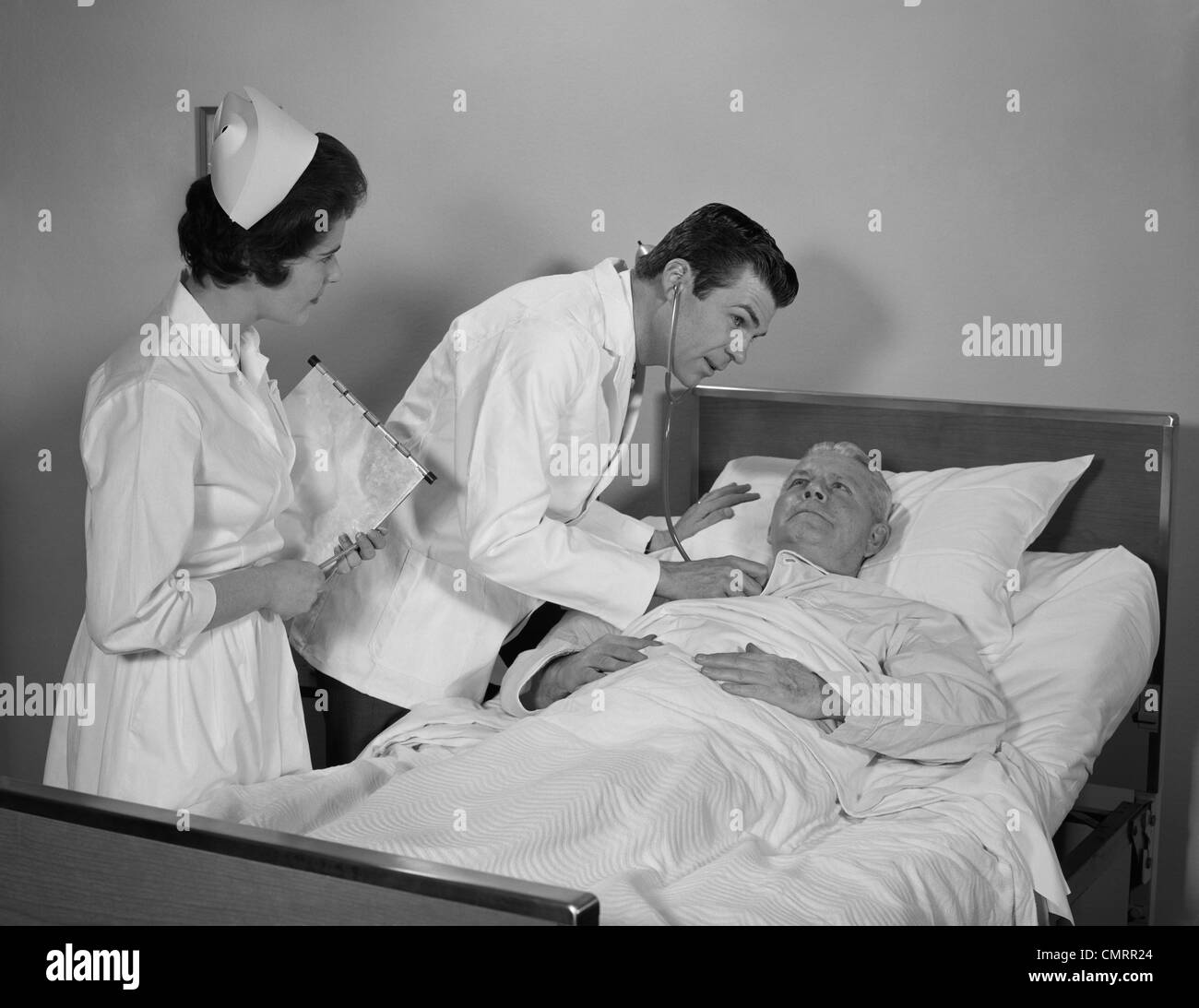 1960s DOCTOR AND NURSE CHECKING ON ELDERLY MALE PATIENT IN HOSPITAL BED Stock Photo