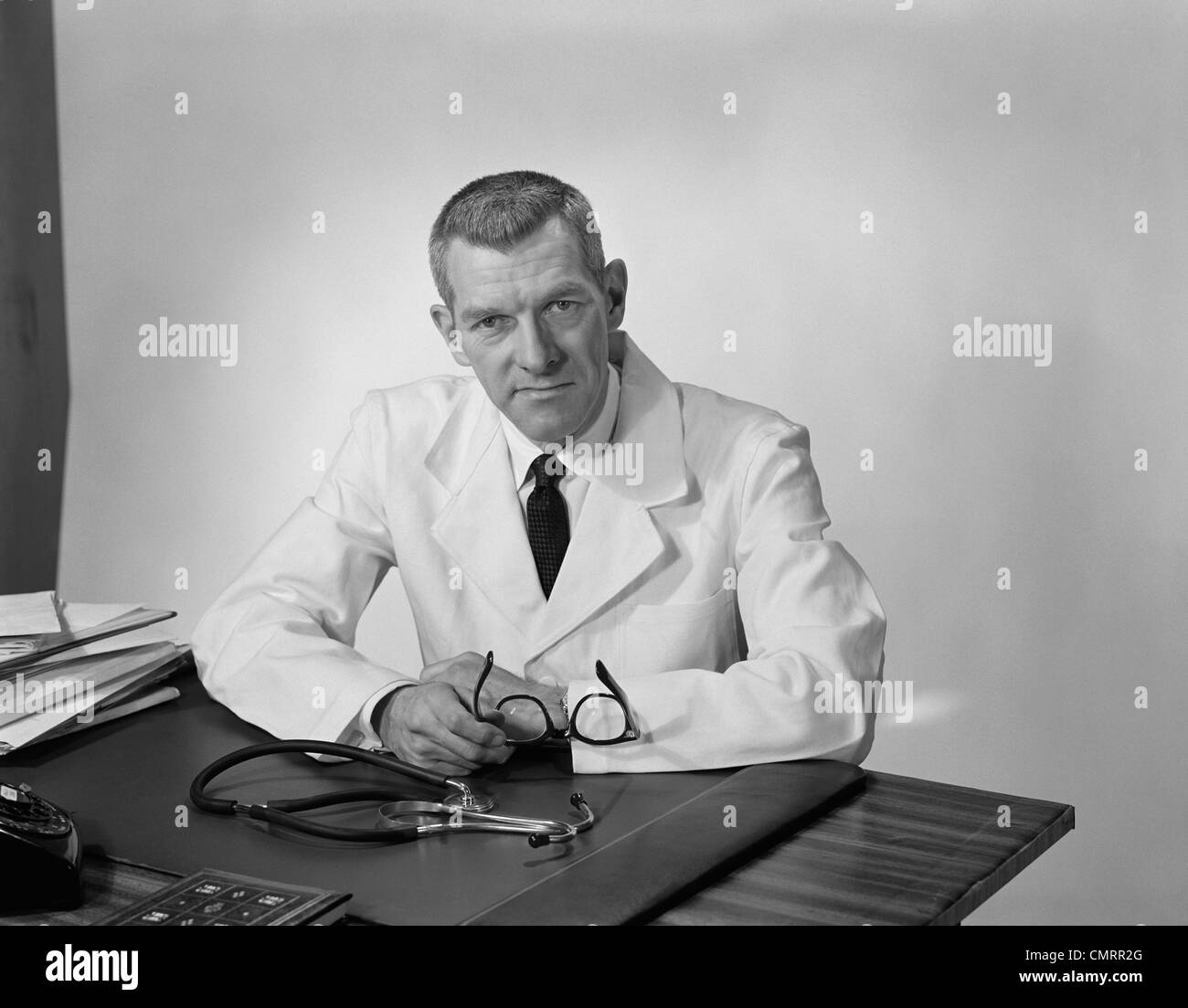 1950s 1960s PORTRAIT MALE DOCTOR SITTING AT DESK SERIOUS EXPRESSION