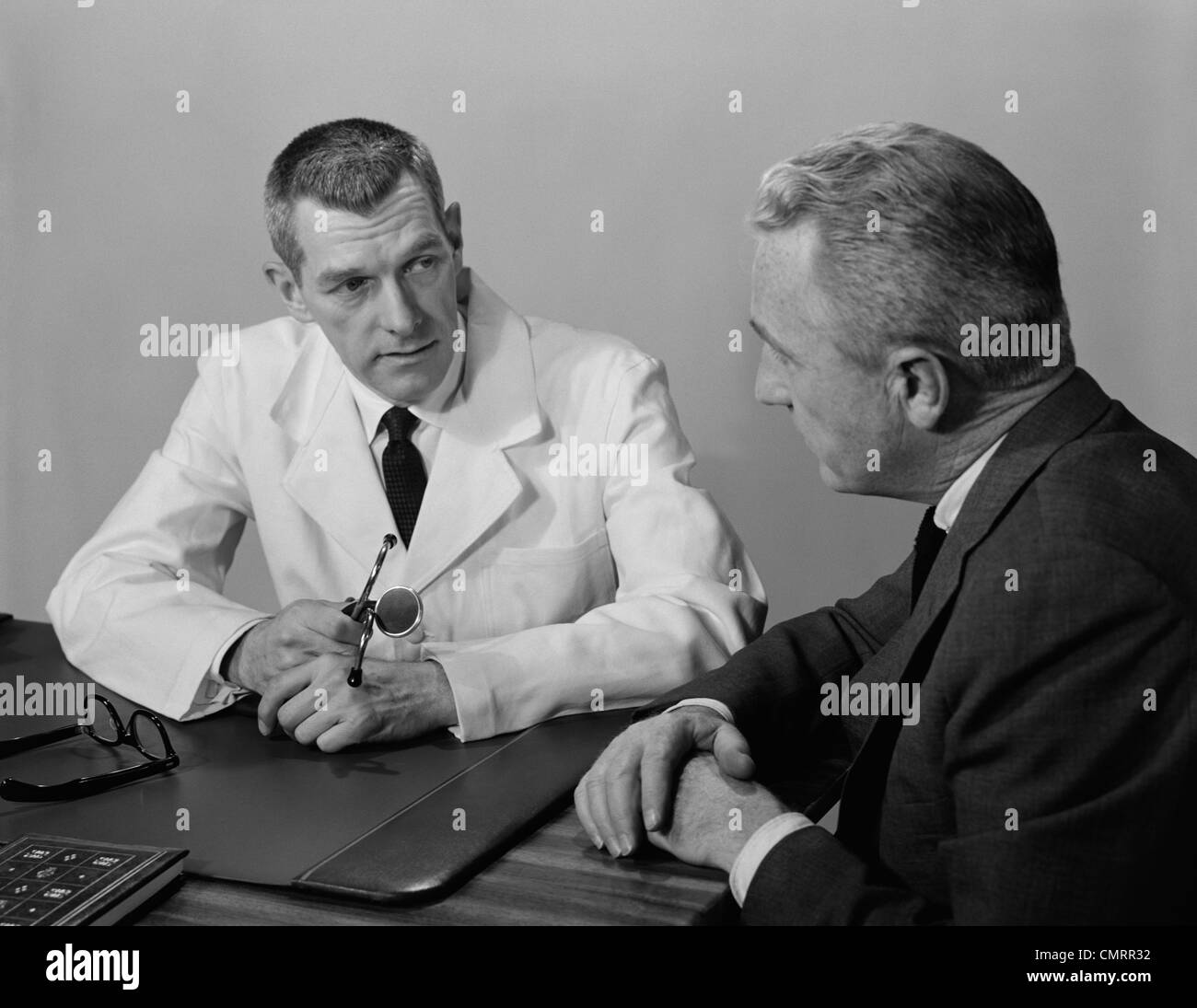 1950s MAN DOCTOR SITTING AT DESK TALKING TO MALE PATIENT Stock Photo