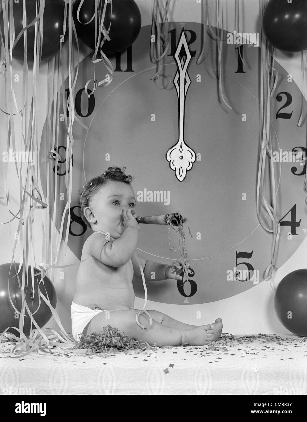 1950s NEW YEAR BABY SITTING BY MIDNIGHT CLOCK FACE BLOWING PARTY NOISE MAKER AMID BALLOONS AND PAPER STREAMERS Stock Photo