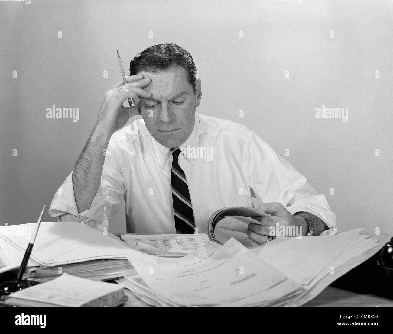 1960s BUSINESSMAN DESK FULL PAPERS HAND TO FOREHEAD SERIOUS EXPRESSION Stock Photo