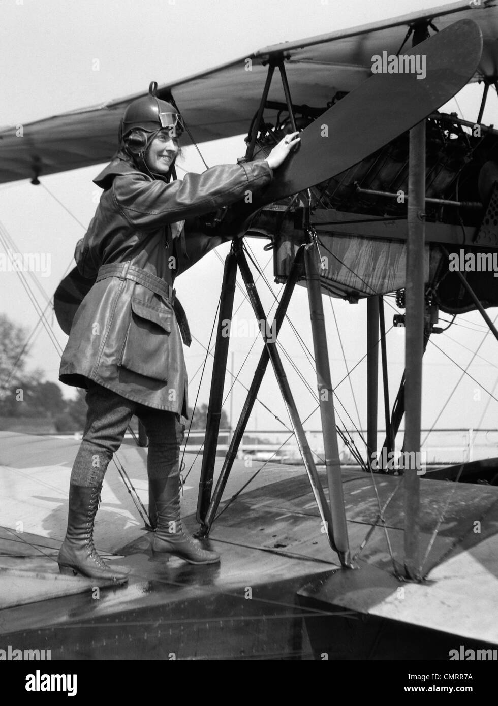 1920s SMILING WOMAN AVIATOR TURNING FLOAT BIPLANE PROPELLER WEARING LEATHER FLYING CLOTHES GOGGLES AND HELMET Stock Photo