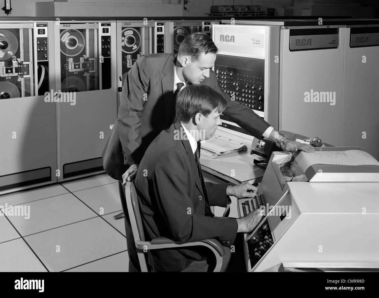 1960s TWO BUSINESSMAN PROGRAMMING LARGE MAINFRAME COMPUTER SURROUNDED BY DATA STORAGE TAPE DRIVES OFFICE INDOOR Stock Photo