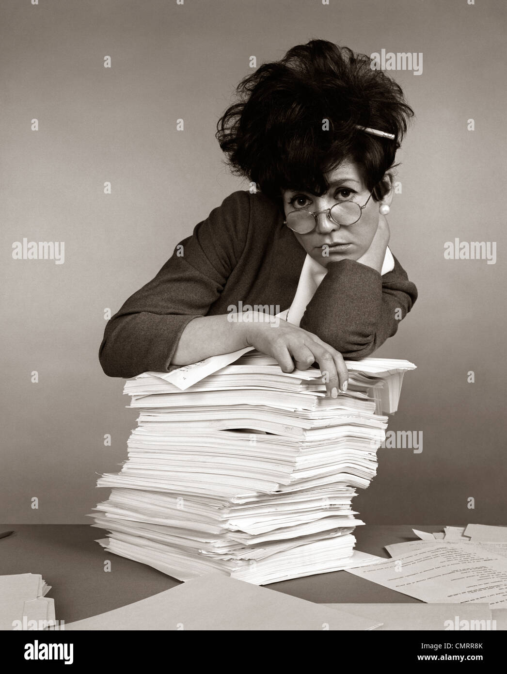1960s 1970s WOMAN SECRETARY PENCIL STUCK IN TEASED HAIR OVERWORKED EXPRESSION LEANING PILE PAPERWORK Stock Photo